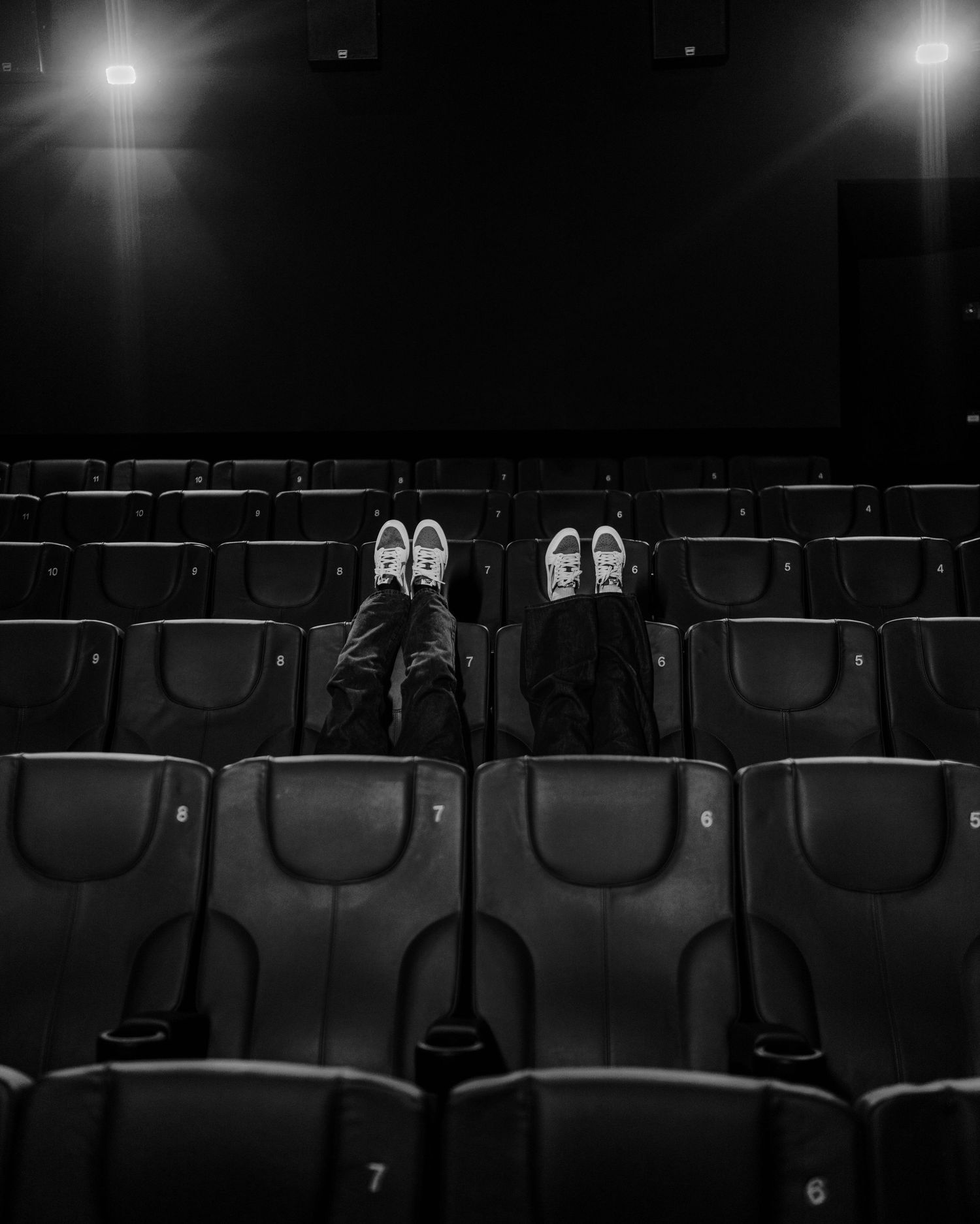 Black and white photo of two people resting feet up on empty cinema seats in a dark movie theater.