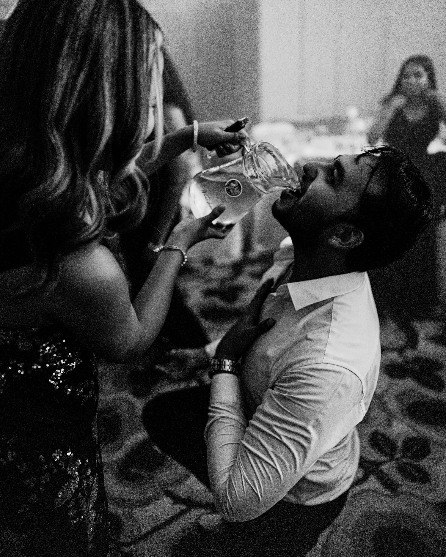 Black and white photo of a woman pouring a drink into a man's mouth at a party event.