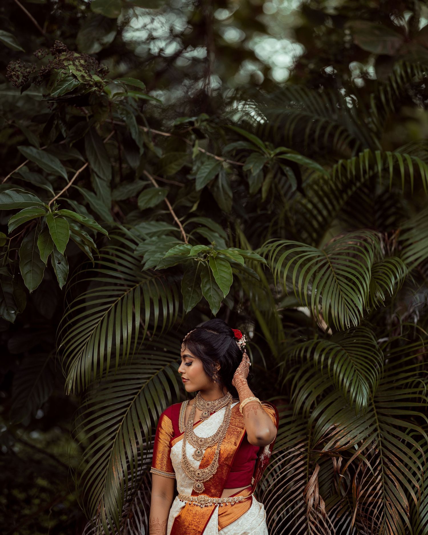 A woman in traditional South Indian bridal attire with gold jewelry stands among lush tropical greenery.