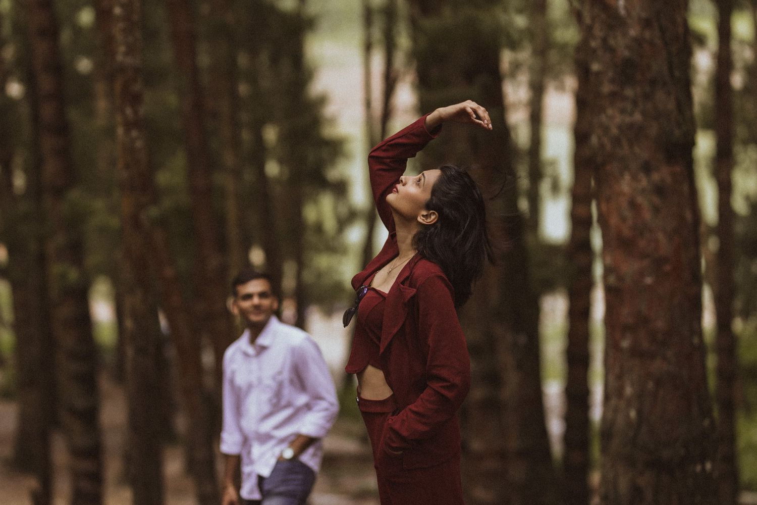 A couple in burgundy and white outfits walk together through a serene forest setting during a photo session.