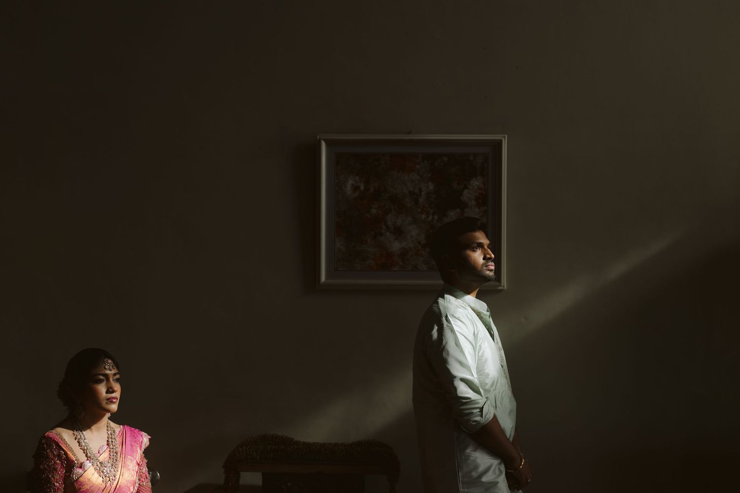 Two figures stand in a dimly lit room with dramatic lighting, creating a moody cinematic atmosphere against dark walls.