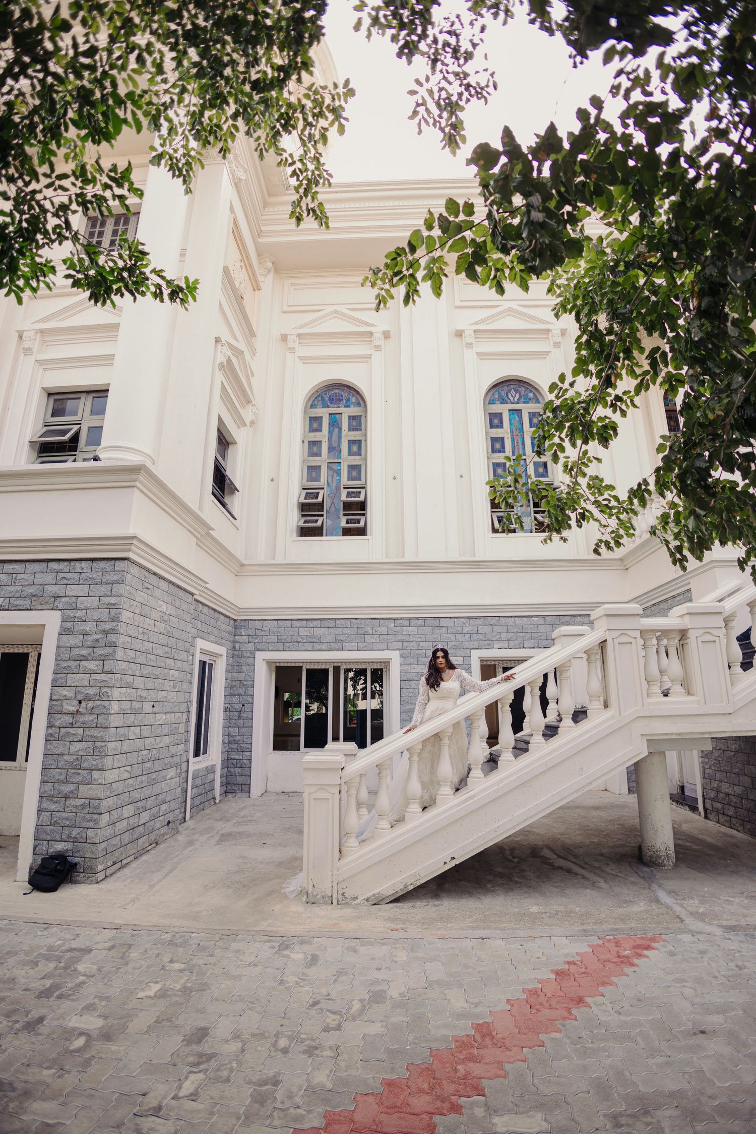 White architectural facade with stone stairs and red carpet runner leading to entrance under leafy trees.