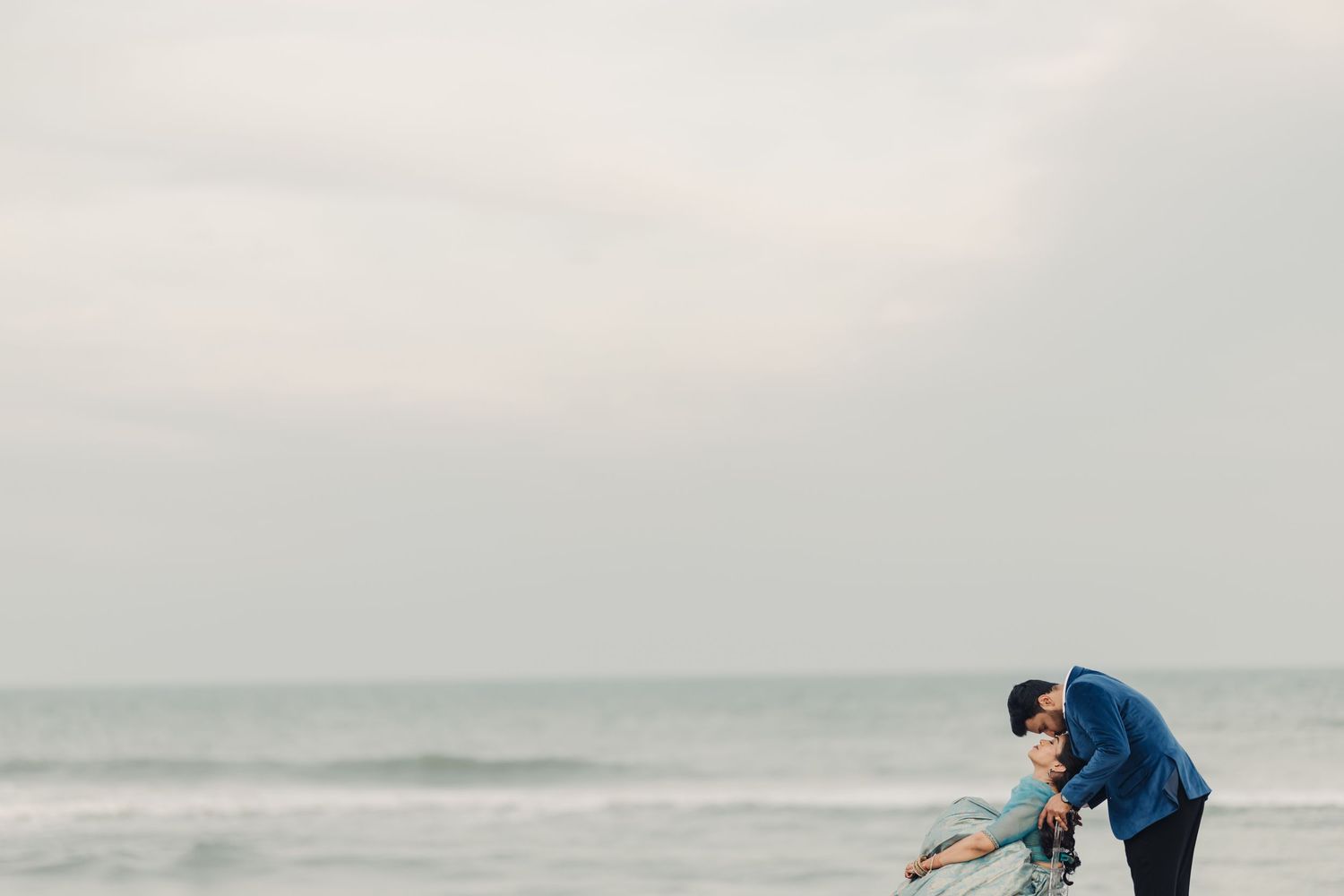 A couple embraces while standing at the edge of a misty beach with gray ocean waves in the background.