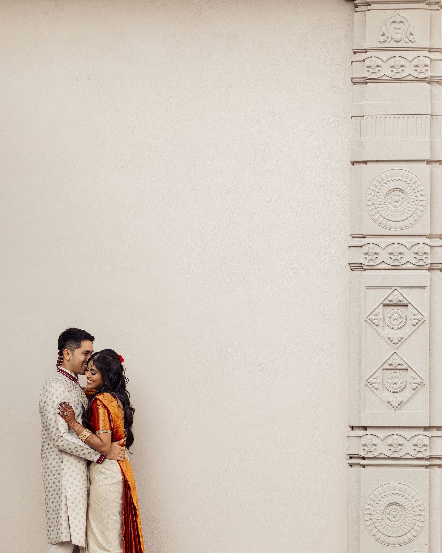 Indian couple in traditional wedding attire embracing tenderly beside an ornate white pillar.
