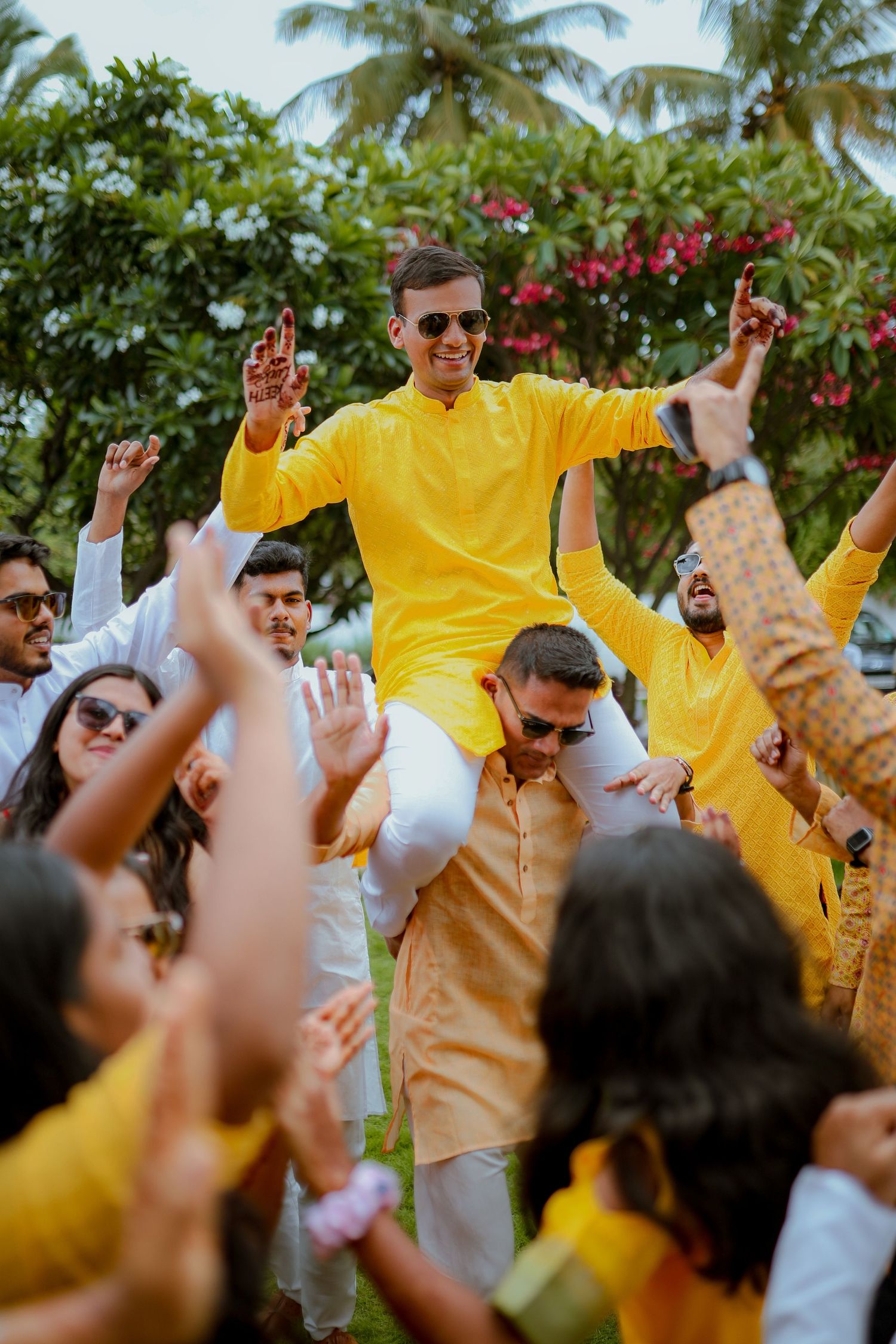 Joyous celebration at an outdoor Indian wedding with guests wearing yellow and white attire surrounded by palm trees and flowers.