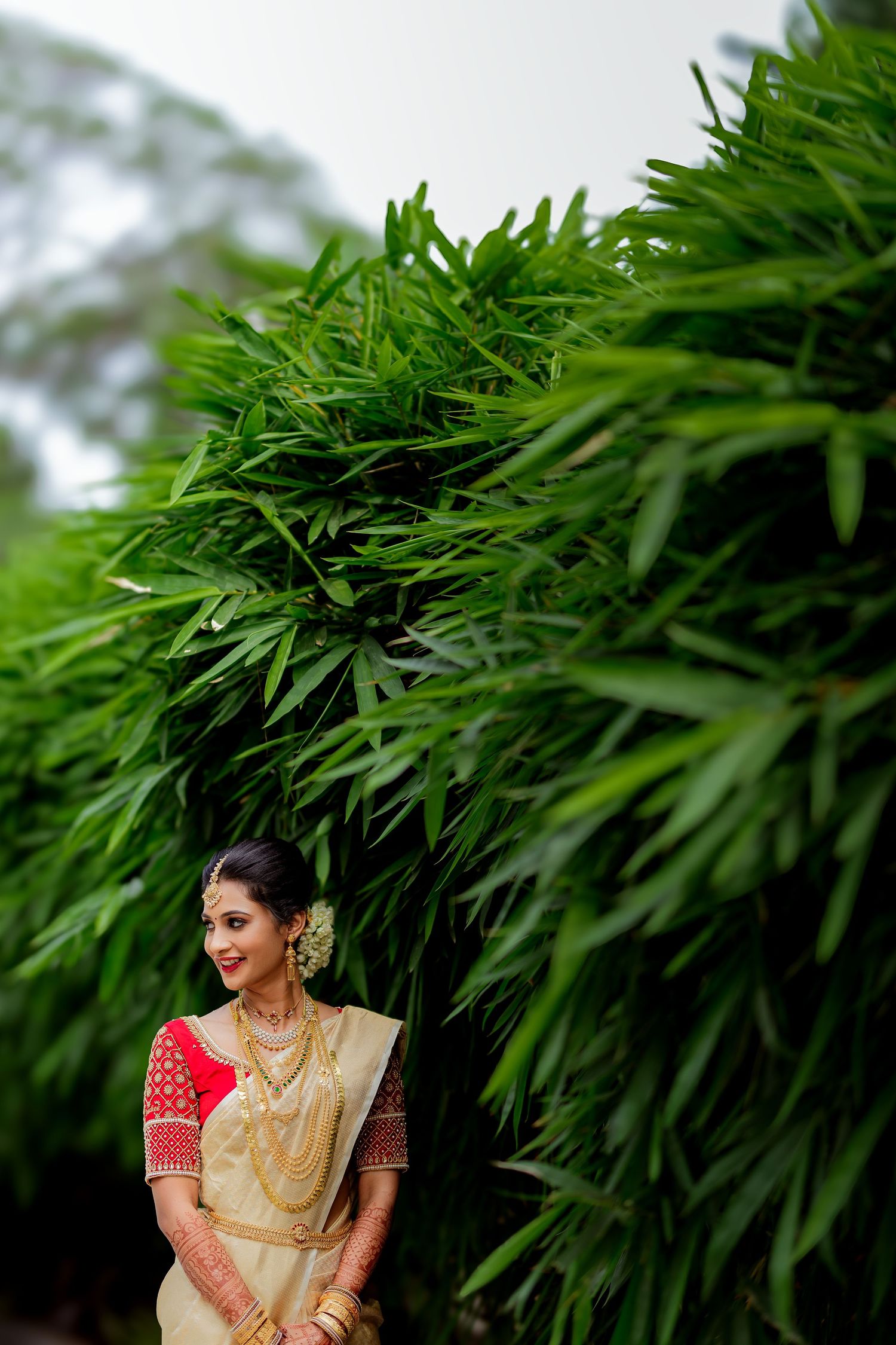 A traditional Indian bride in a beige silk saree and red blouse poses elegantly against lush green palm fronds.