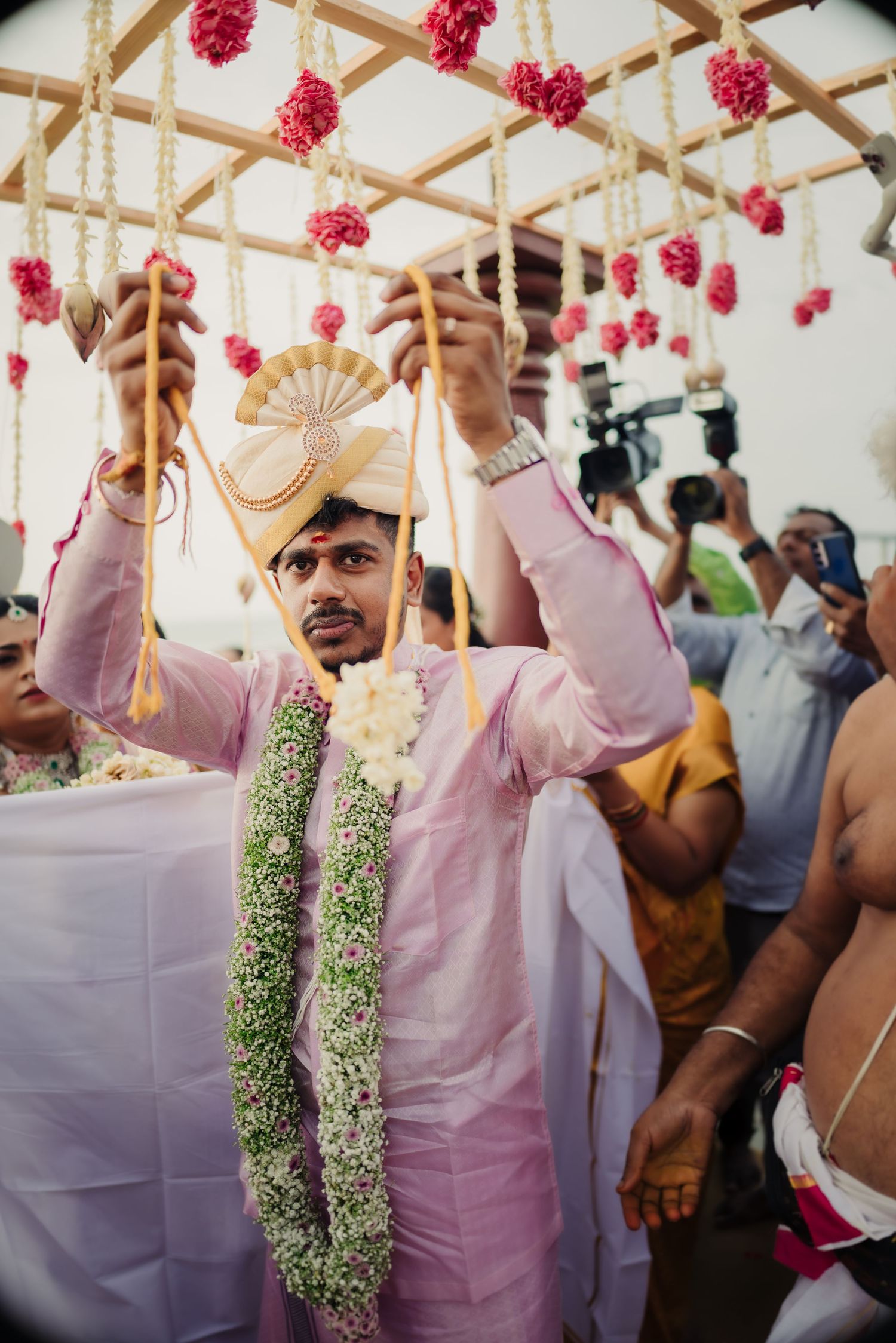 People gather under a decorated ceiling with pink flowers and hanging garlands at a festive celebration.
