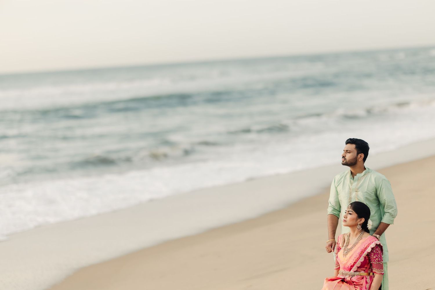 Two people walking together on a sandy beach with ocean waves and cloudy skies.