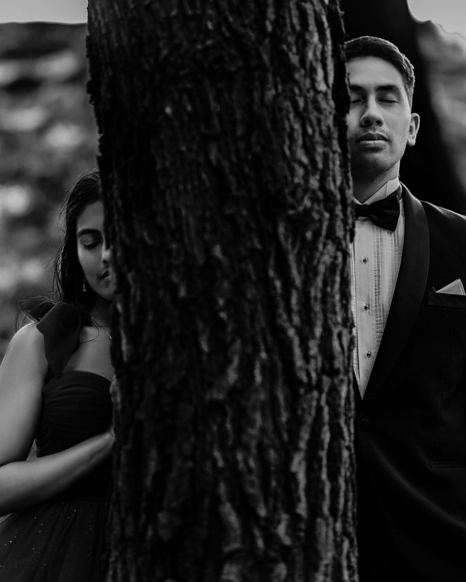Black and white photo of a couple posing on opposite sides of a tree, man in tuxedo, woman in elegant dress.