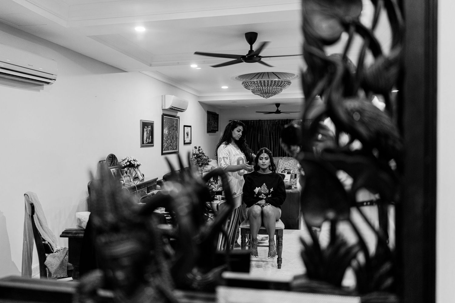 Black and white photo of women getting ready indoors, reflected in a mirror surrounded by tropical plants.