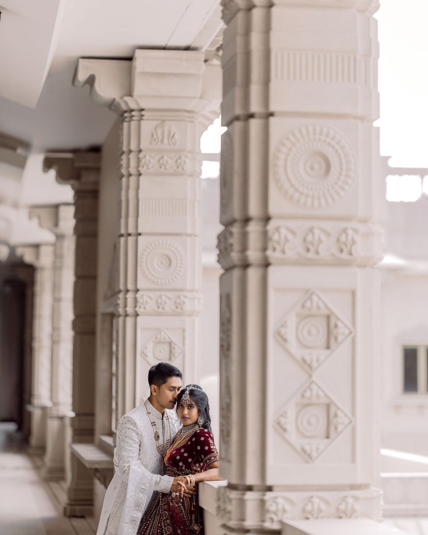 Couple in traditional South Asian wedding attire posing between ornate white carved columns at a temple.