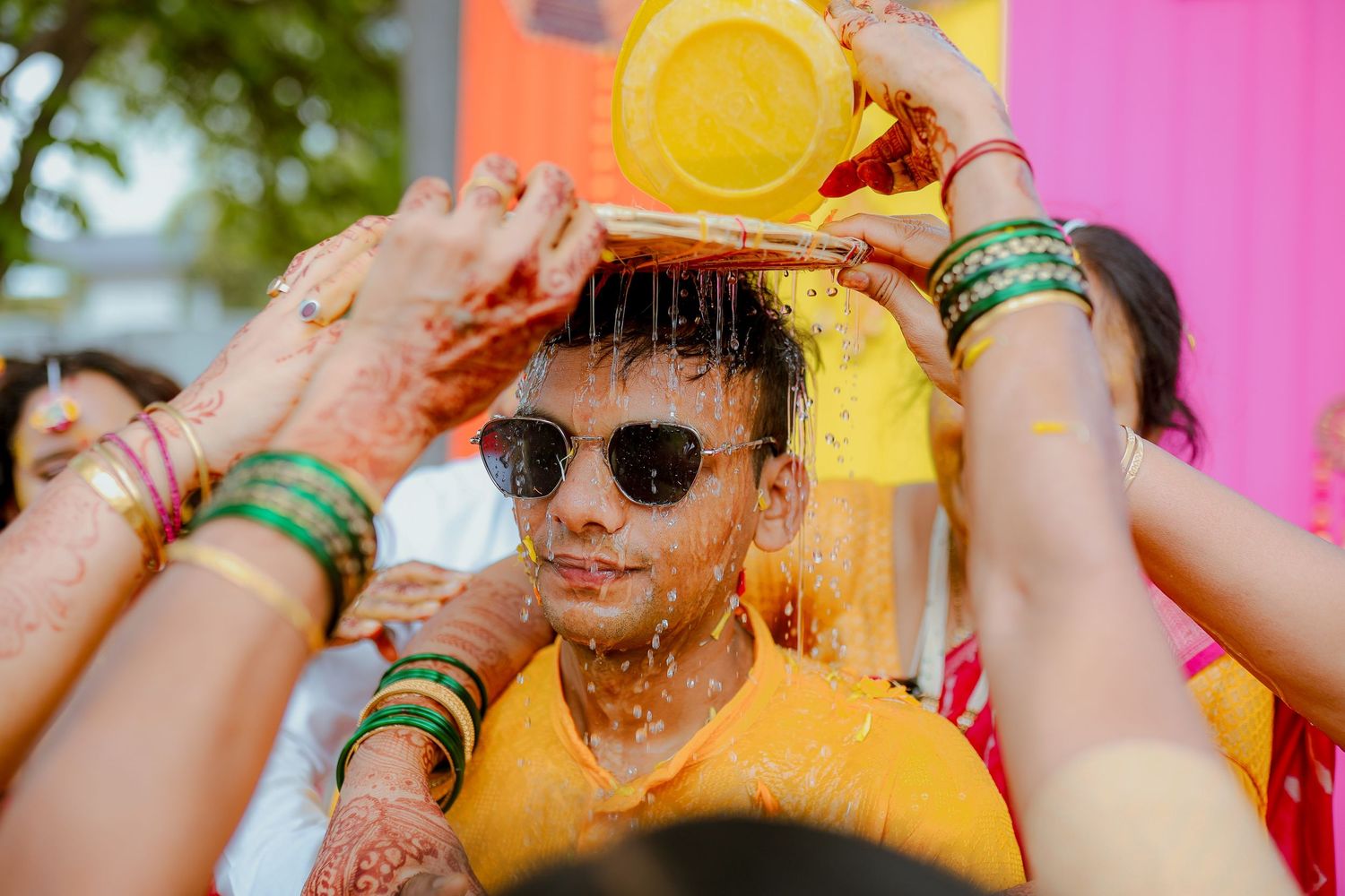 People celebrating Holi festival with yellow powder and colorful bangles against vibrant pink and orange background.
