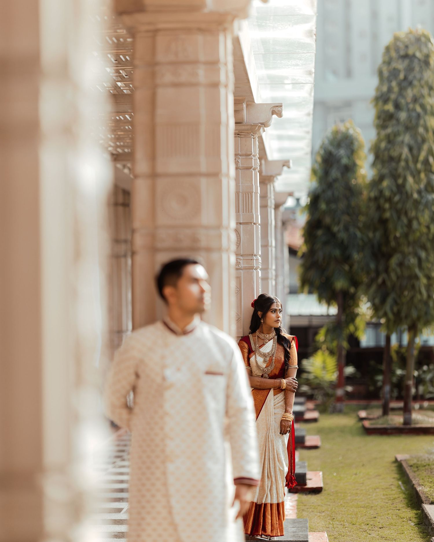 Indian couple in traditional wedding attire posing between ornate columns, bride in gold-bordered saree, groom in white kurta.