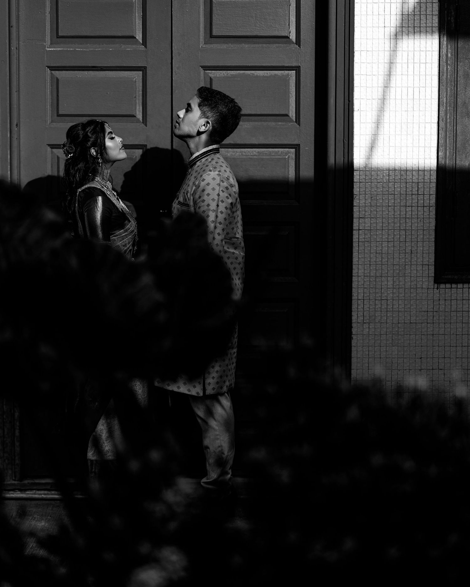 Black and white photo of a couple sharing a romantic moment near ornate wooden doors in traditional Indian attire.