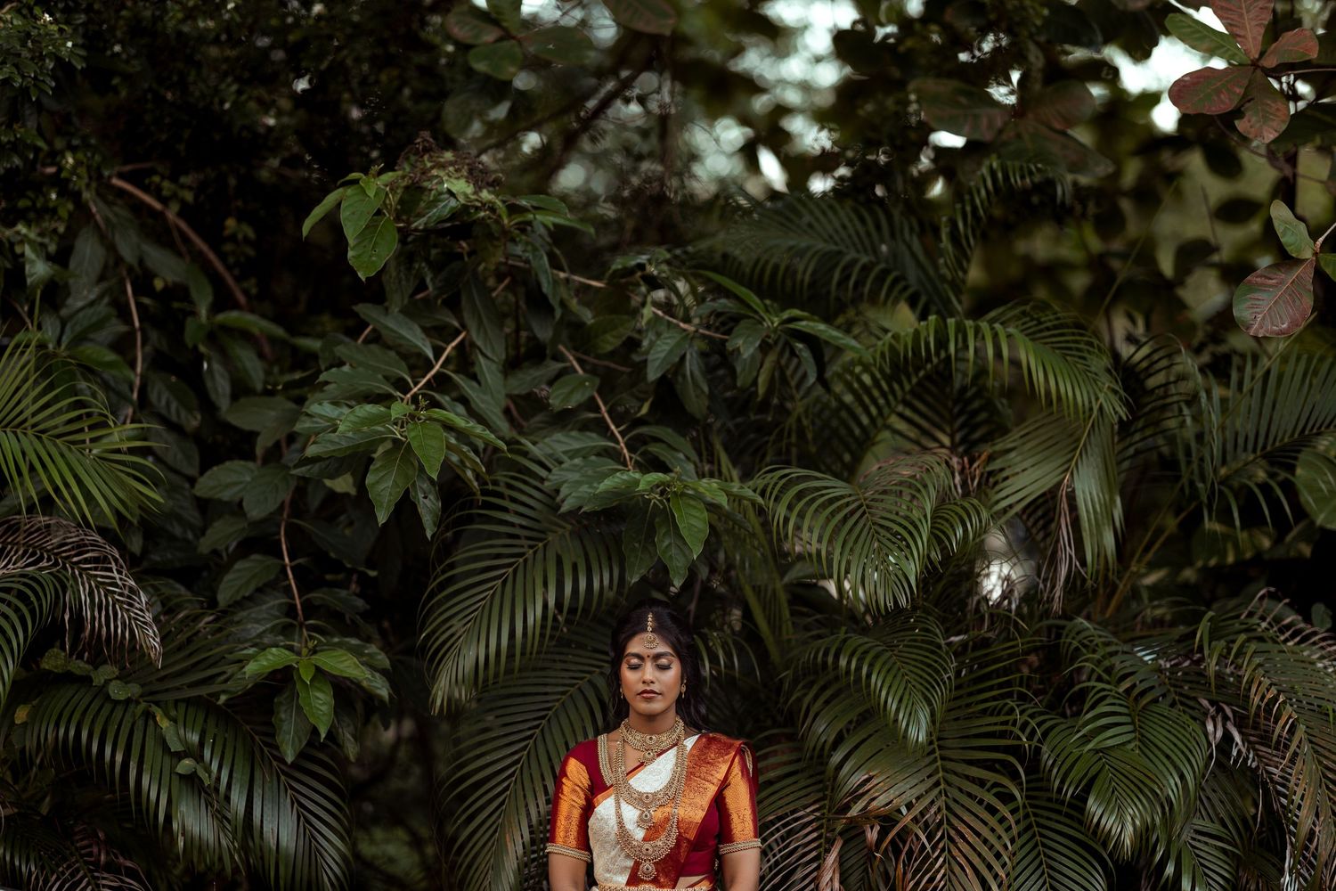Woman in traditional Indian saree meditating peacefully surrounded by lush tropical green foliage.