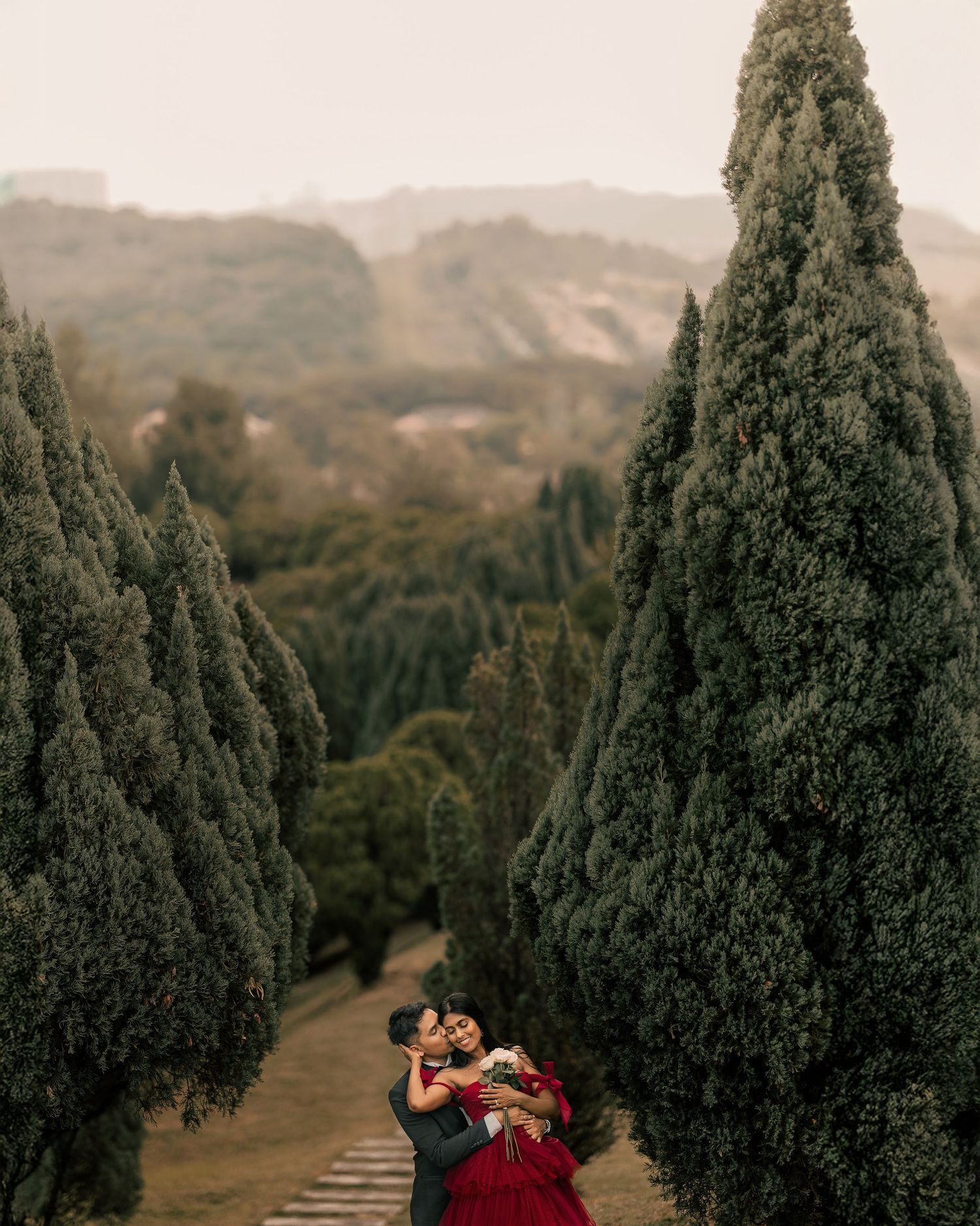 Couple embracing romantically between tall cypress trees with misty hills in the background.