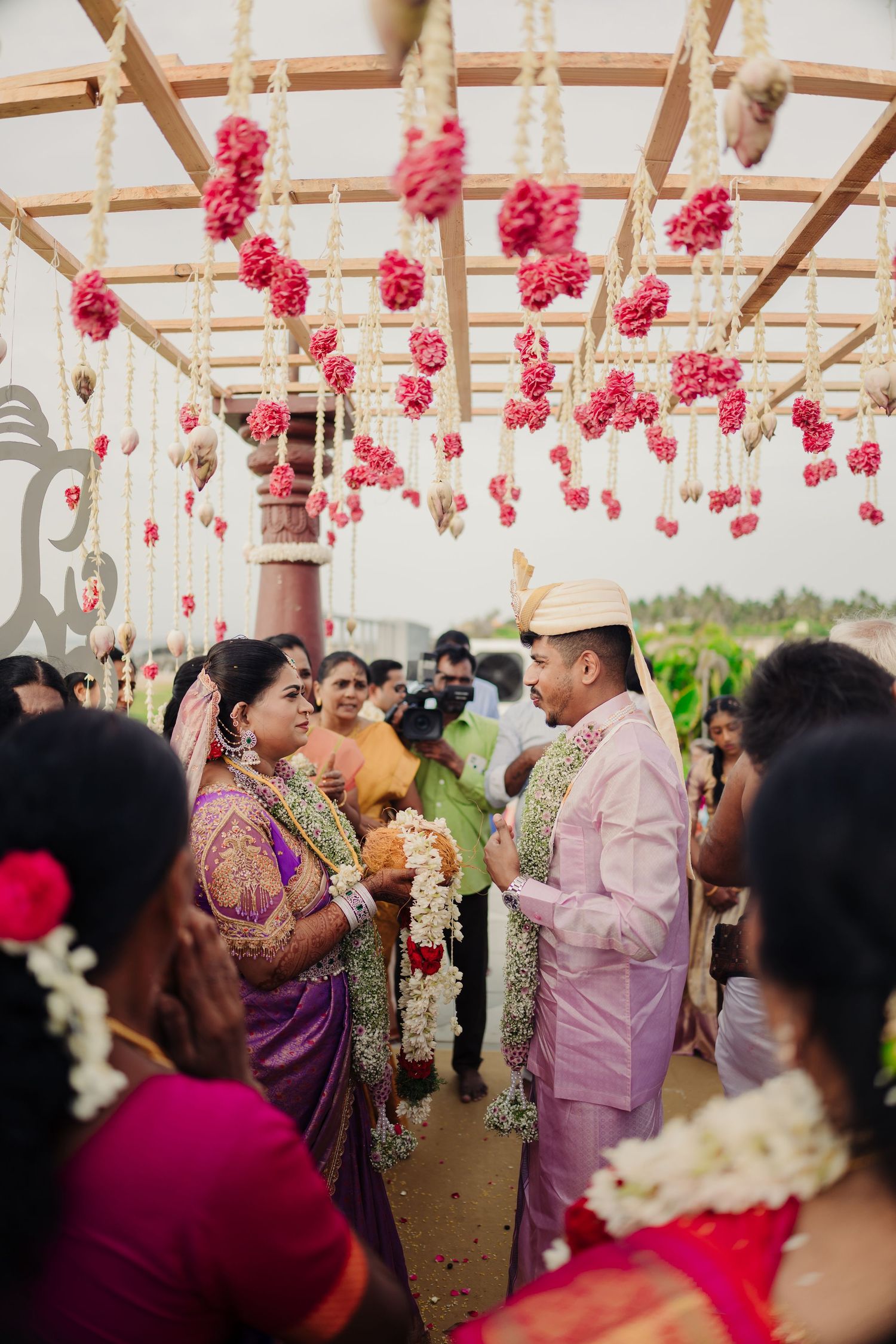Traditional Indian wedding ceremony under a canopy decorated with hanging pink floral arrangements and guests gathered around.