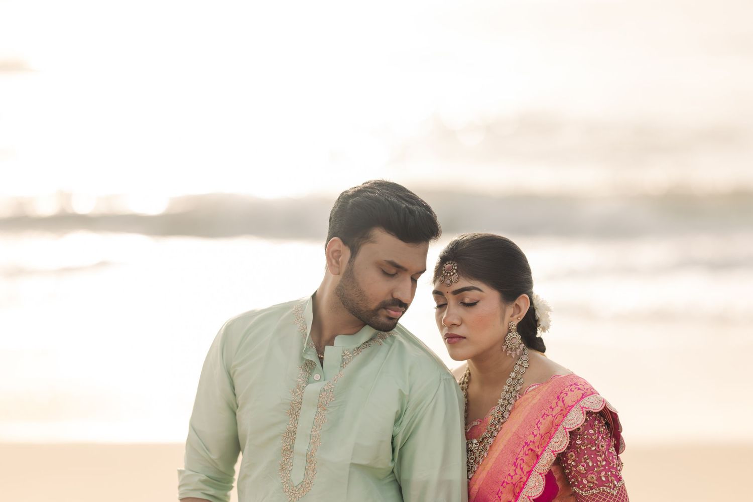 Intimate moment between couple in traditional Indian dress sitting on beach at sunset.