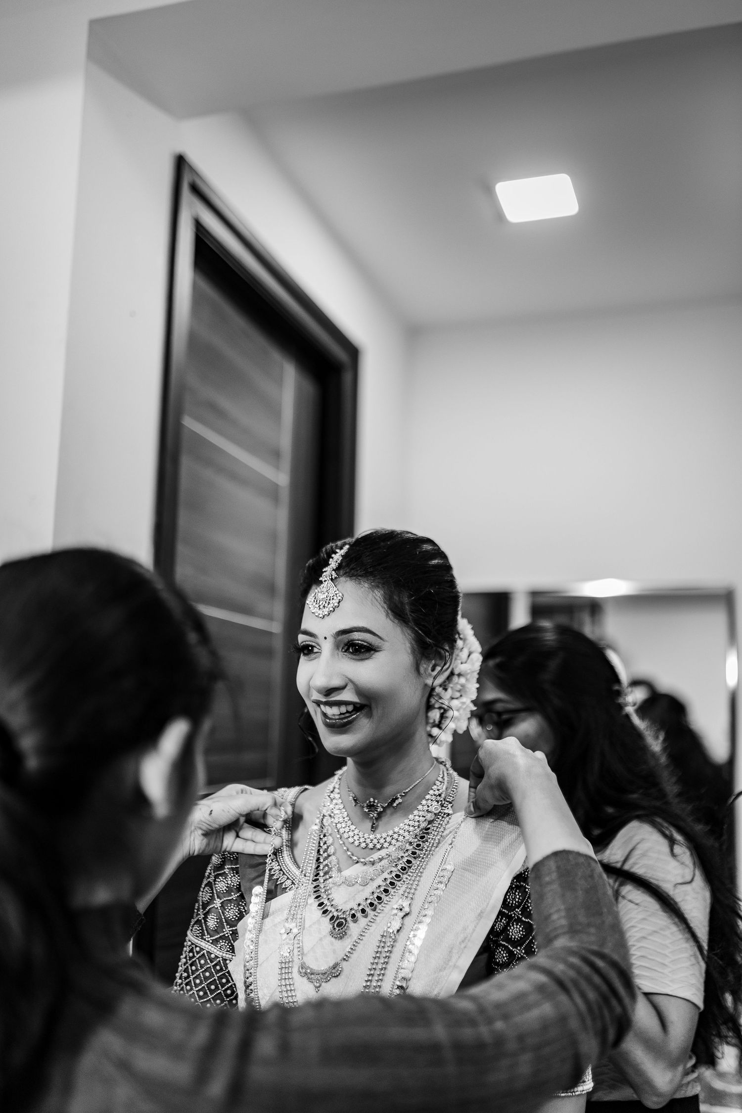 A black and white candid photo shows a joyful moment during an Indian wedding celebration with traditional jewelry.