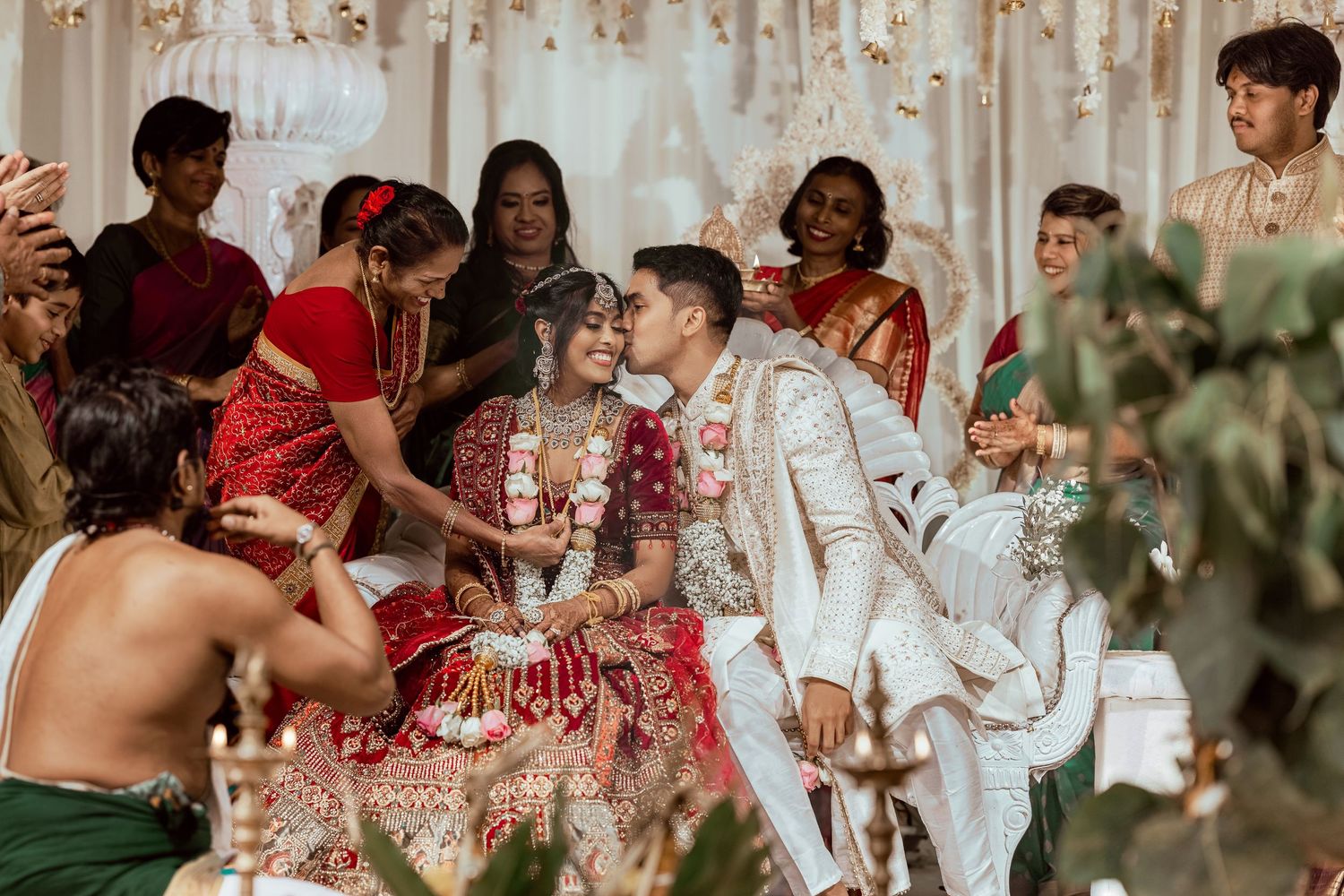 Indian bride and groom share a kiss during traditional Hindu wedding ceremony surrounded by guests in colorful traditional attire.