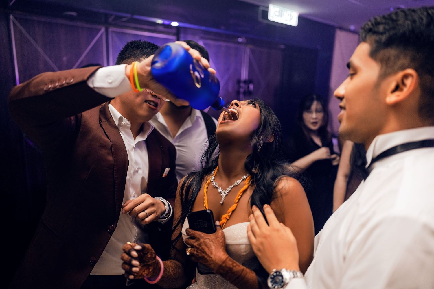 People partying at a nightclub event, with a man pouring a blue bottle drink into a woman's mouth on the dance floor.