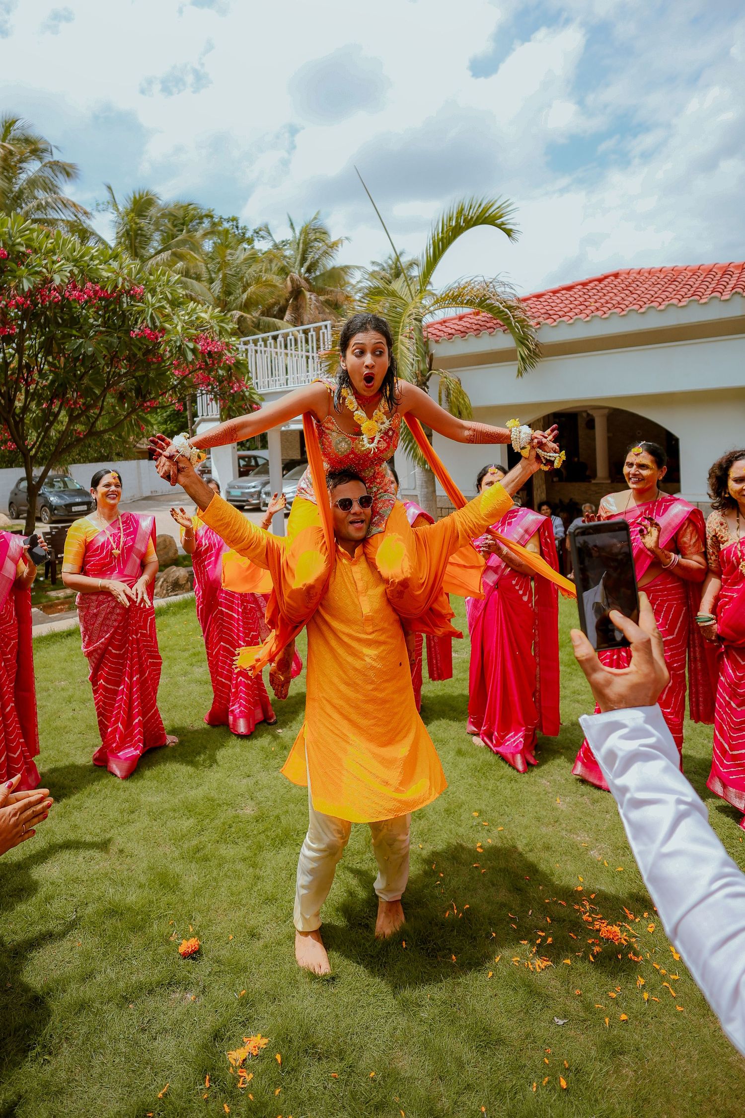 A lively outdoor celebration with dancers in vibrant red and orange traditional attire performing on a green lawn.