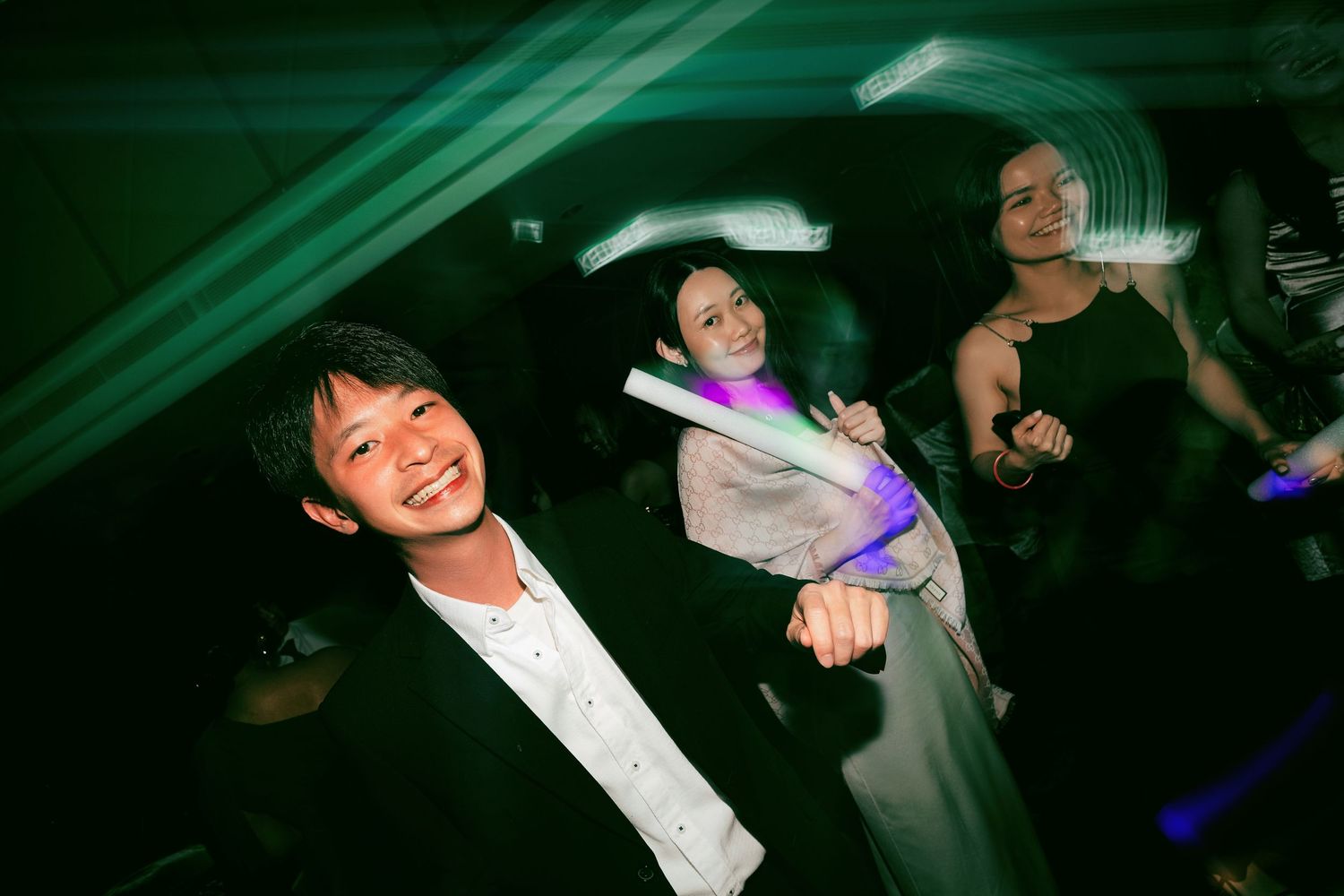 Three people dancing and smiling at a party, holding glow sticks under green lights.