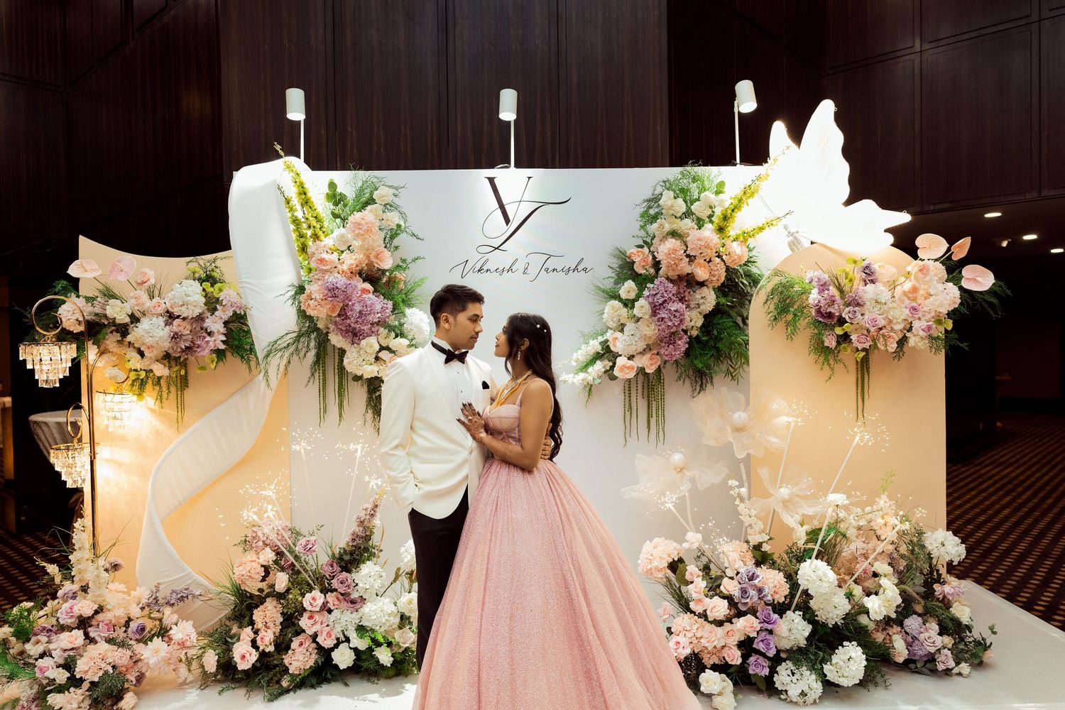 Couple in formal attire poses before an elegant floral wedding backdrop with pink and white blooms.