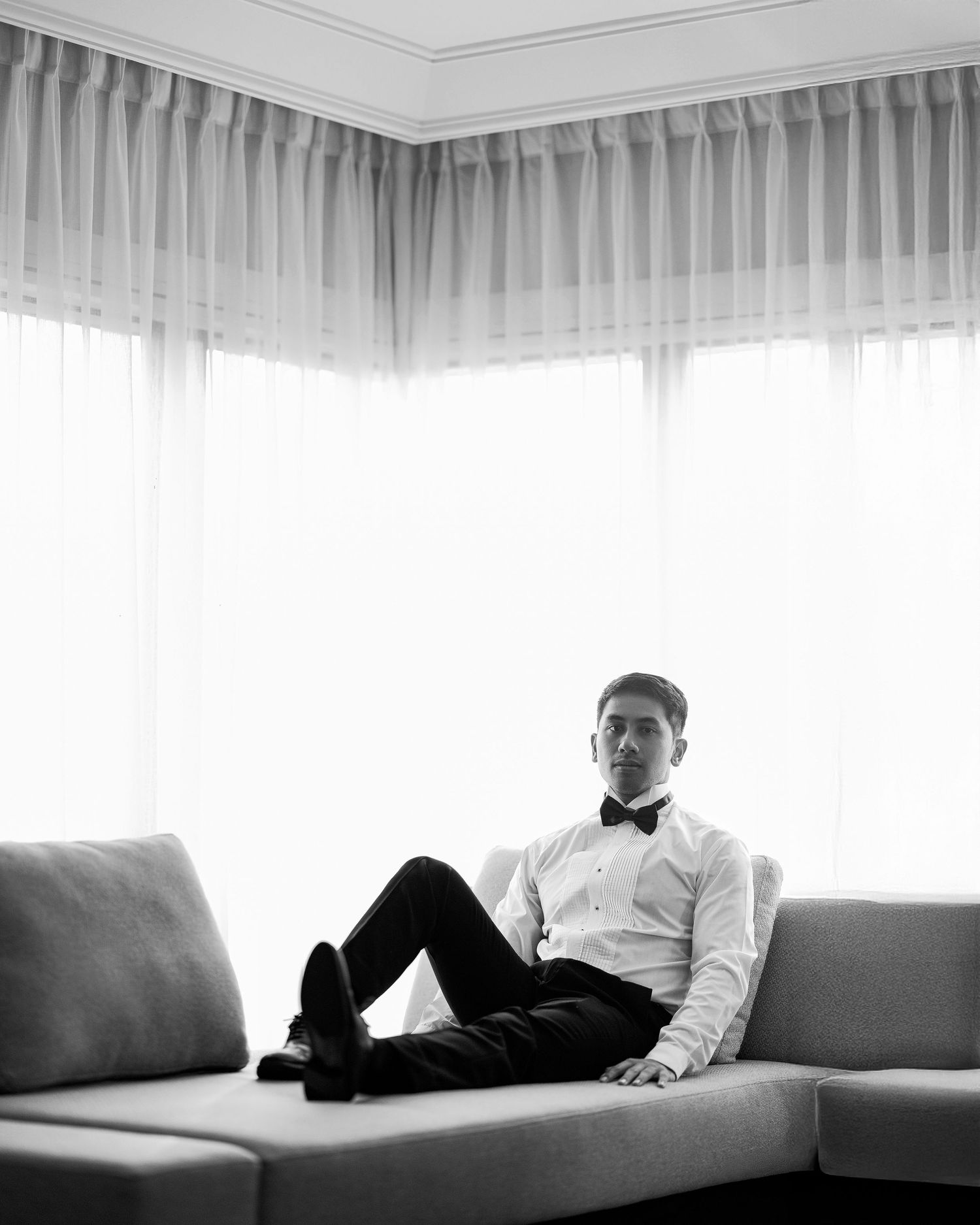 A man in a tuxedo and bow tie reclines elegantly on a modern sofa in a black and white portrait photo.