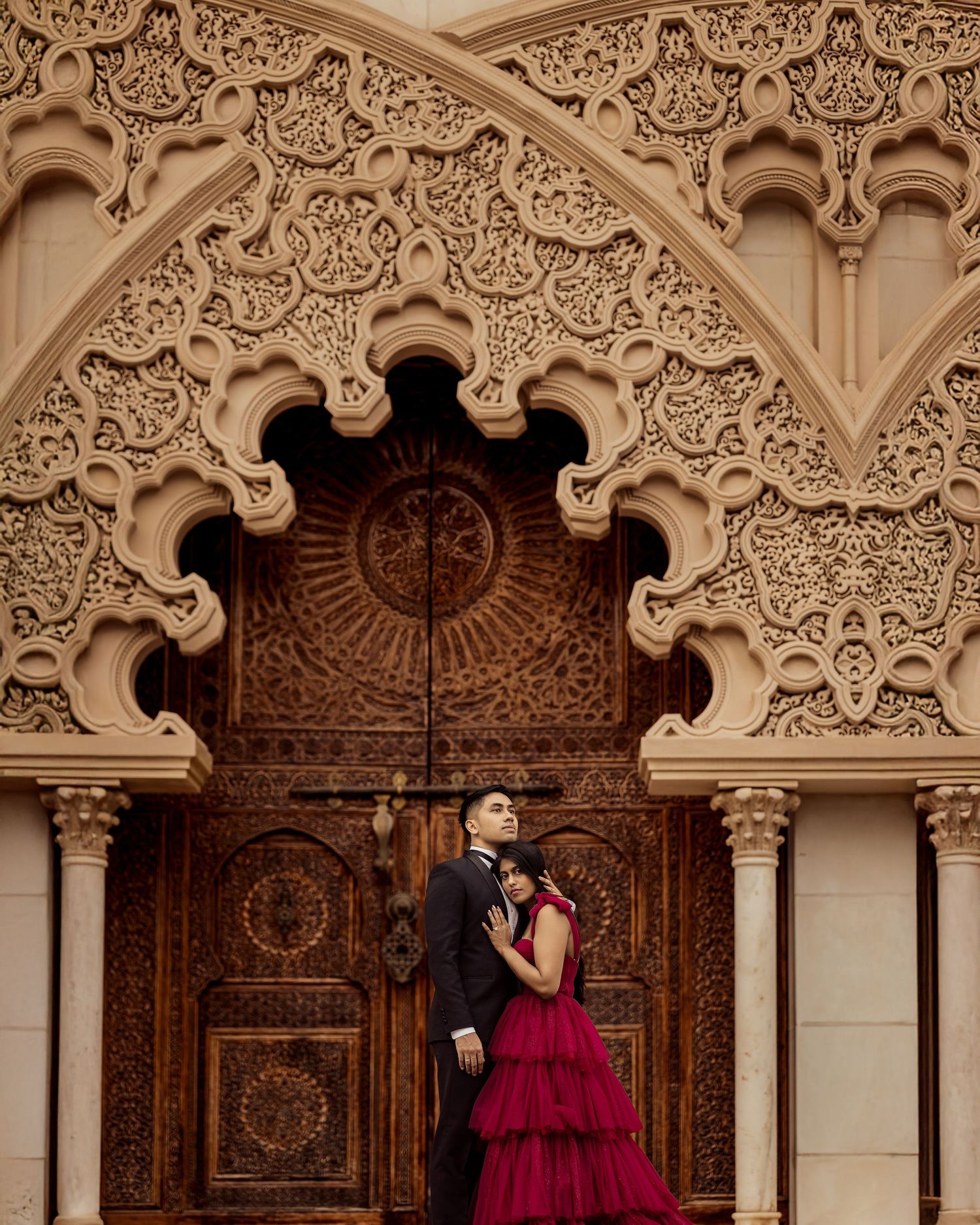 Couple in formal attire poses in front of ornate Moorish-style architecture with detailed carved arches and columns.
