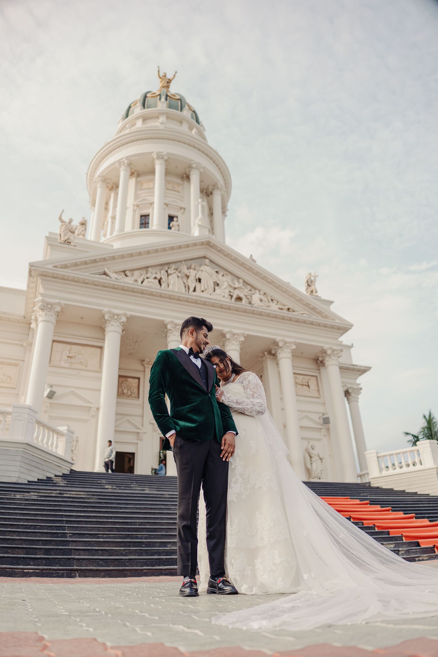 Elegant wedding couple poses on grand white stone steps of a classical domed building with red carpet.