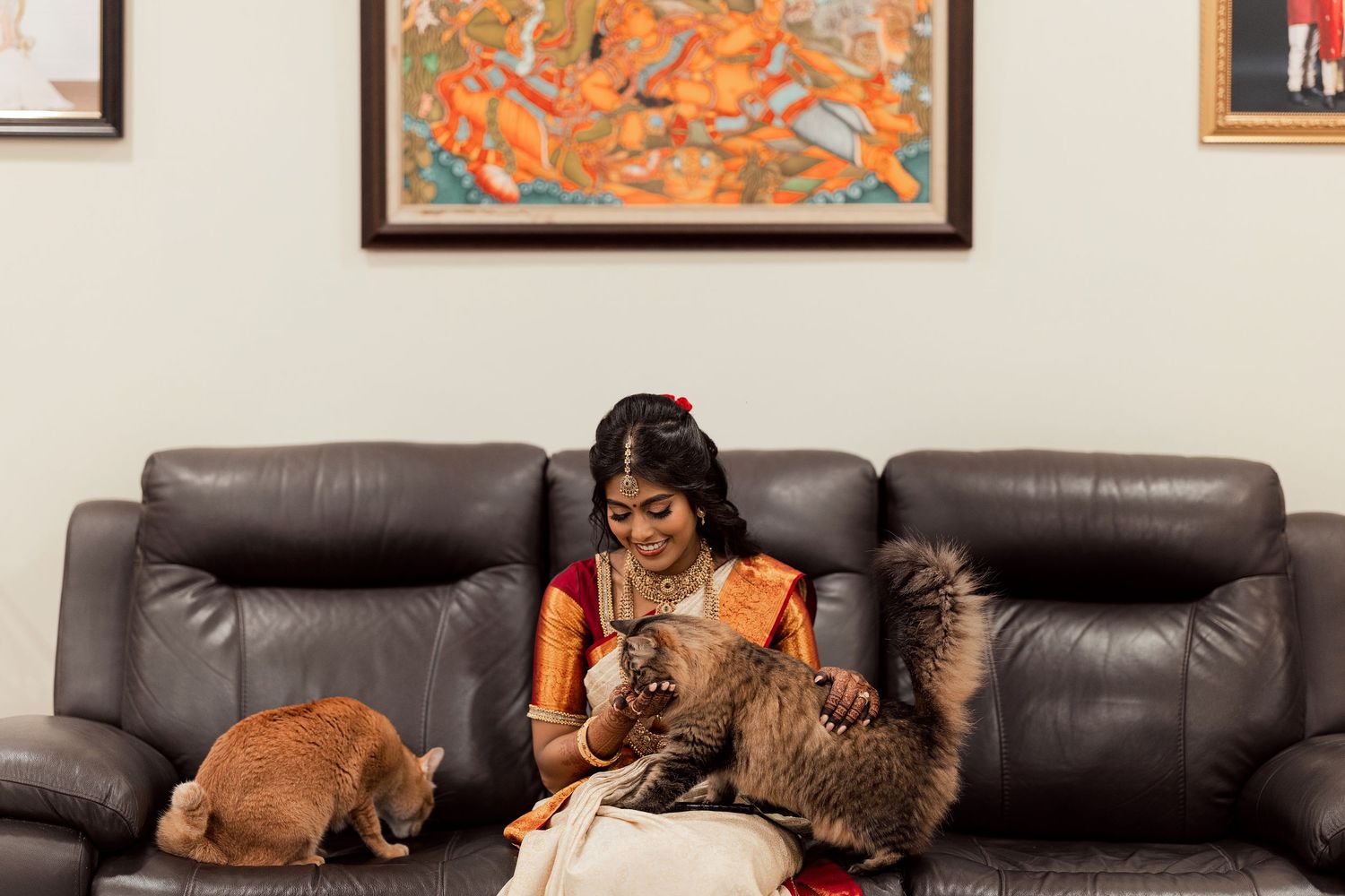 A woman in a traditional Indian saree sits on a gray leather sofa, gently holding a fluffy cat while another cat rests nearby.