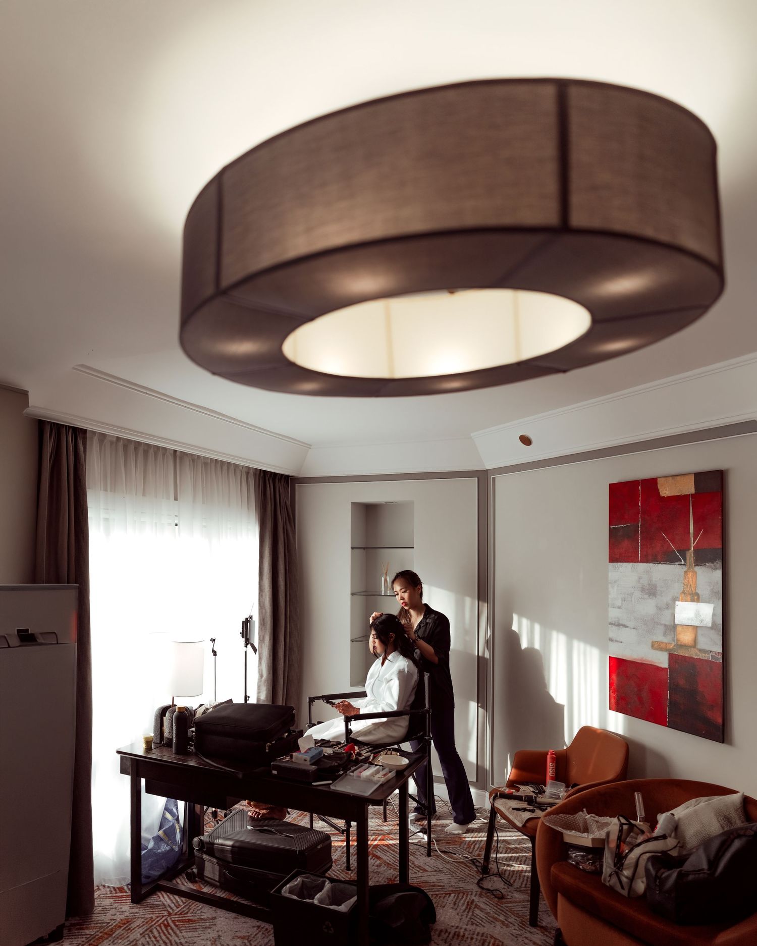 Hairstylist working on a client in a modern hotel room with elegant drum ceiling light and contemporary decor.