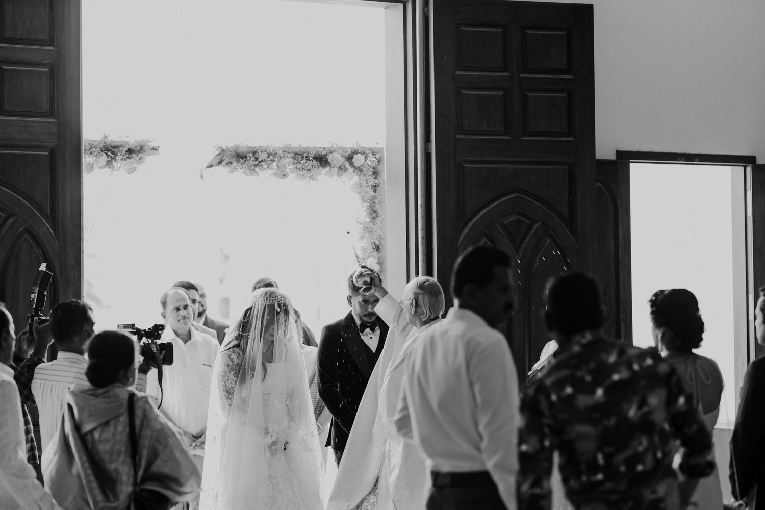 A black and white photo shows a group of people in formal attire standing in silhouette against a bright window.