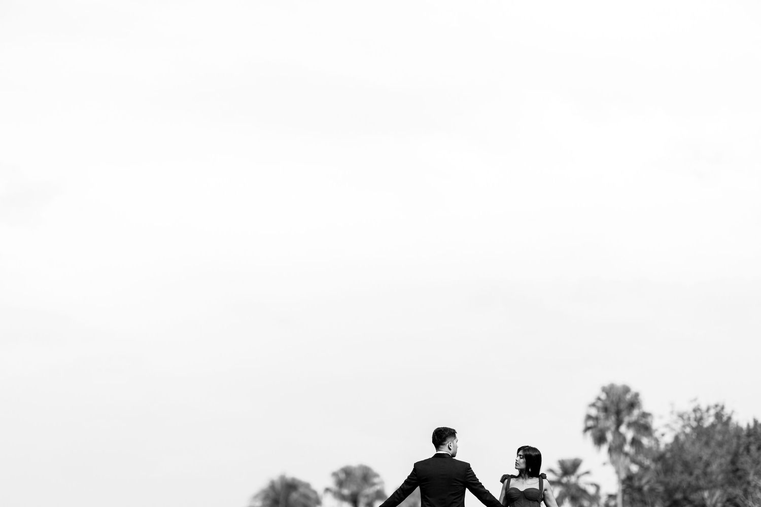 Black and white photo of a couple holding hands walking toward palm trees under a bright sky.