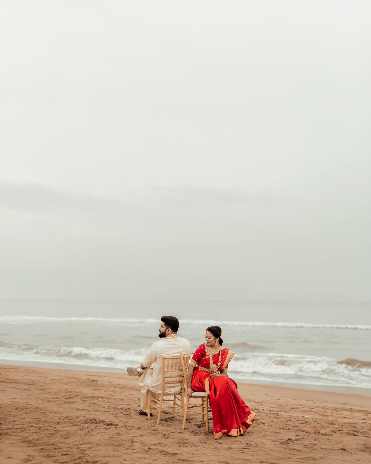 Two people in red attire sit in wooden chairs on a misty beach looking out at the ocean waves.