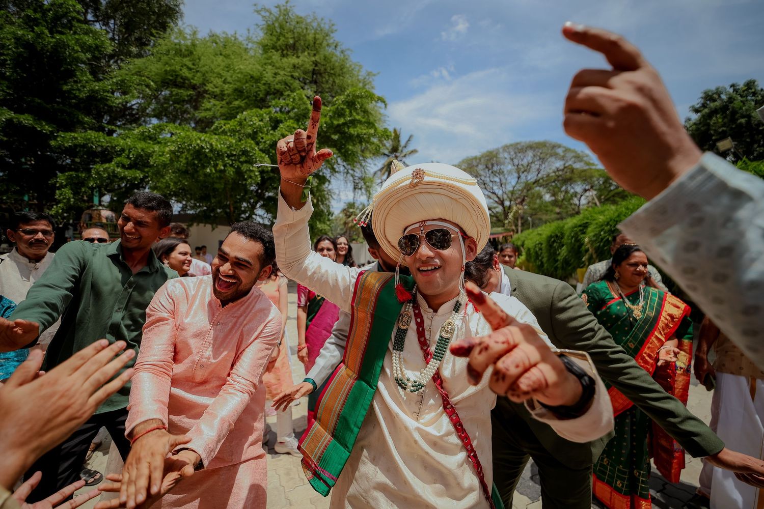 Wedding guests dance and celebrate outdoors on a sunny day with joyful expressions and raised arms.