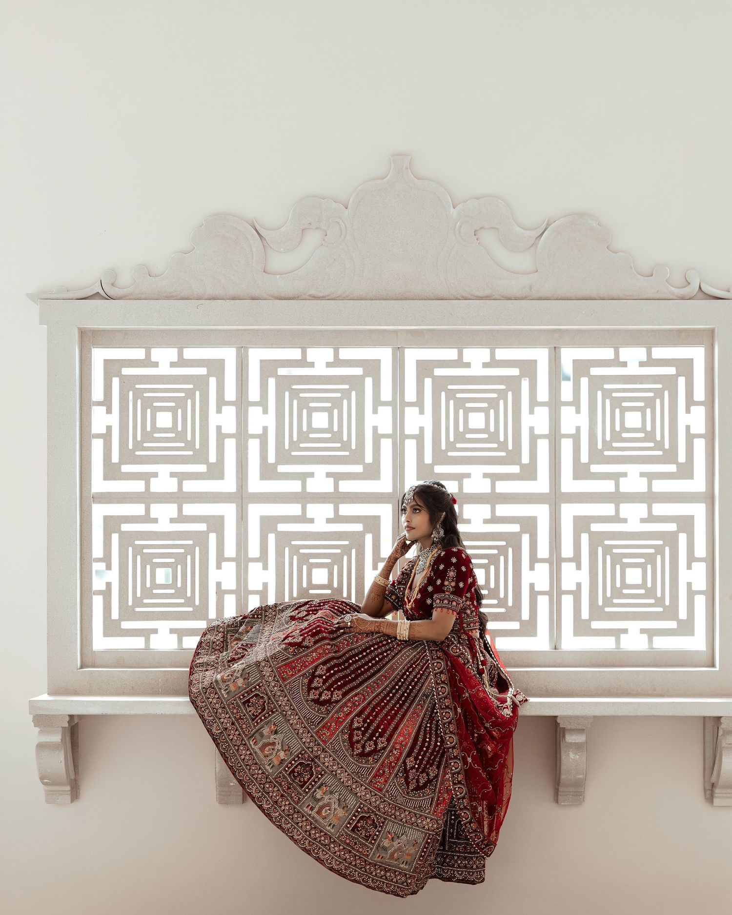 A bride in a red embroidered lehenga sits gracefully against an ornate white lattice panel backdrop.