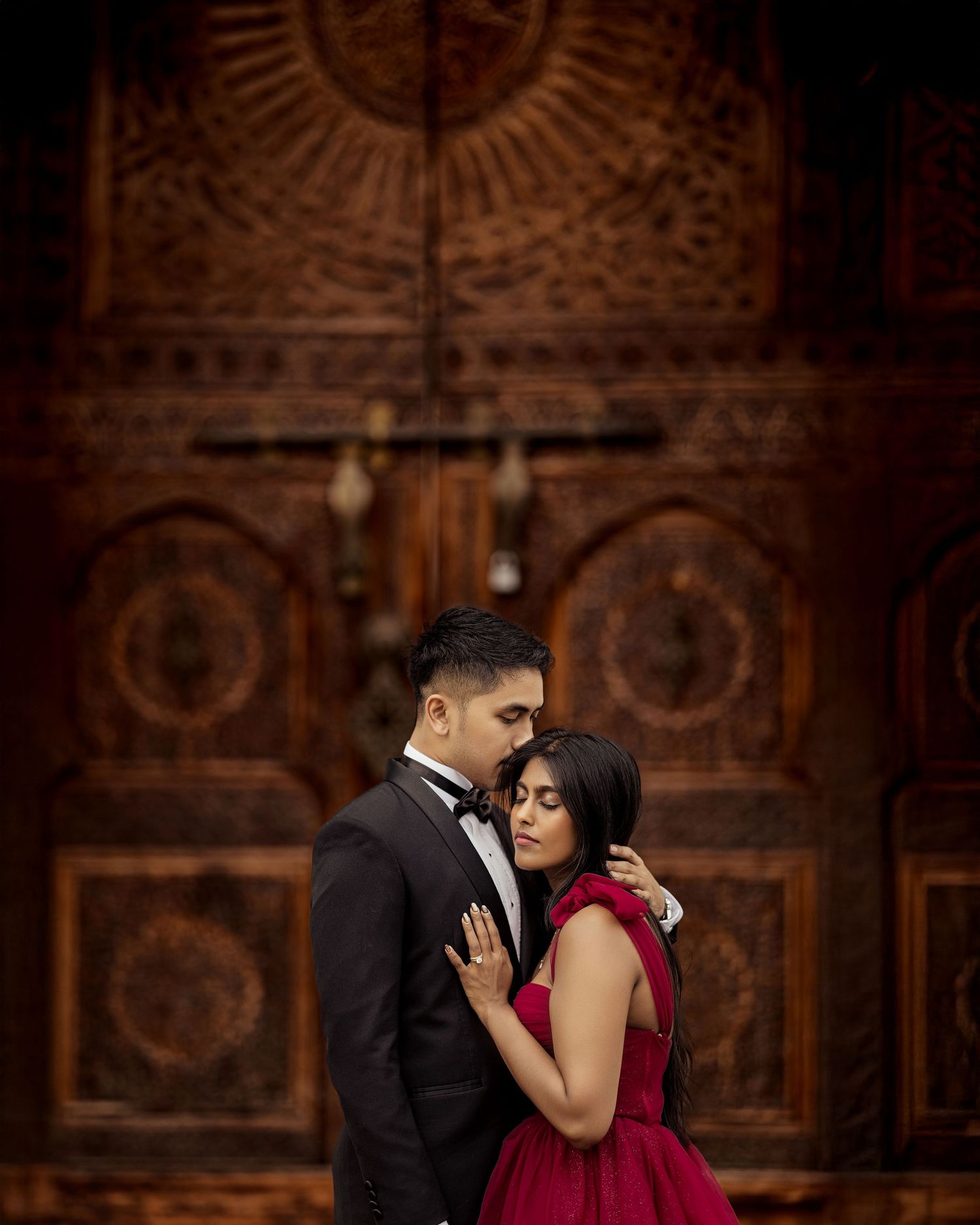 A romantic couple in formal attire embrace in front of an ornate wooden wall with decorative carvings.