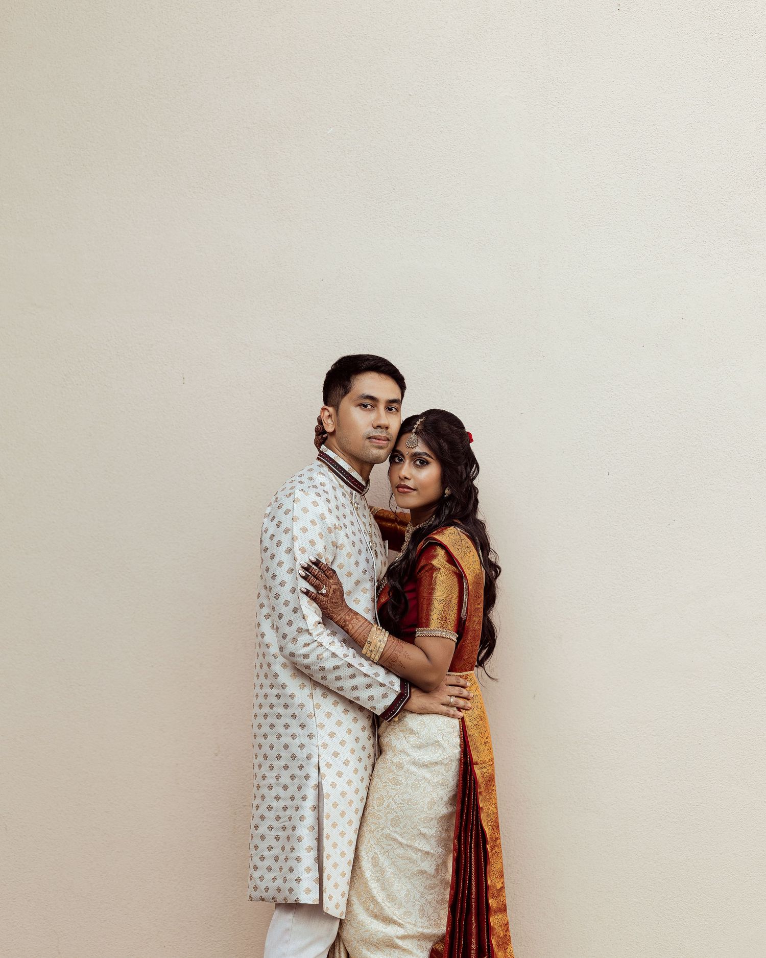 Indian couple in traditional attire embracing, man in white sherwani and woman in gold and red saree, against neutral wall.