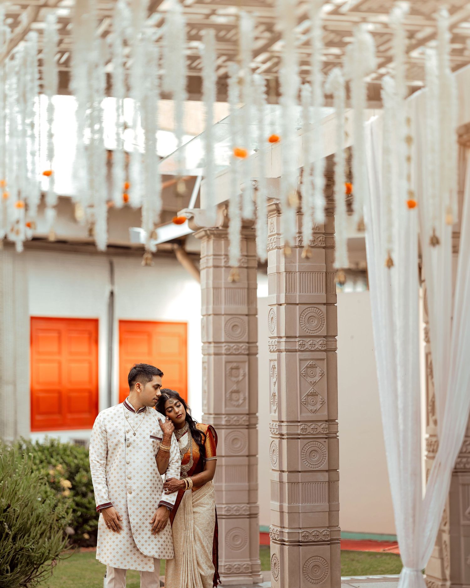 Indian couple in traditional wedding attire standing between ornate pillars under floral white draping decor.