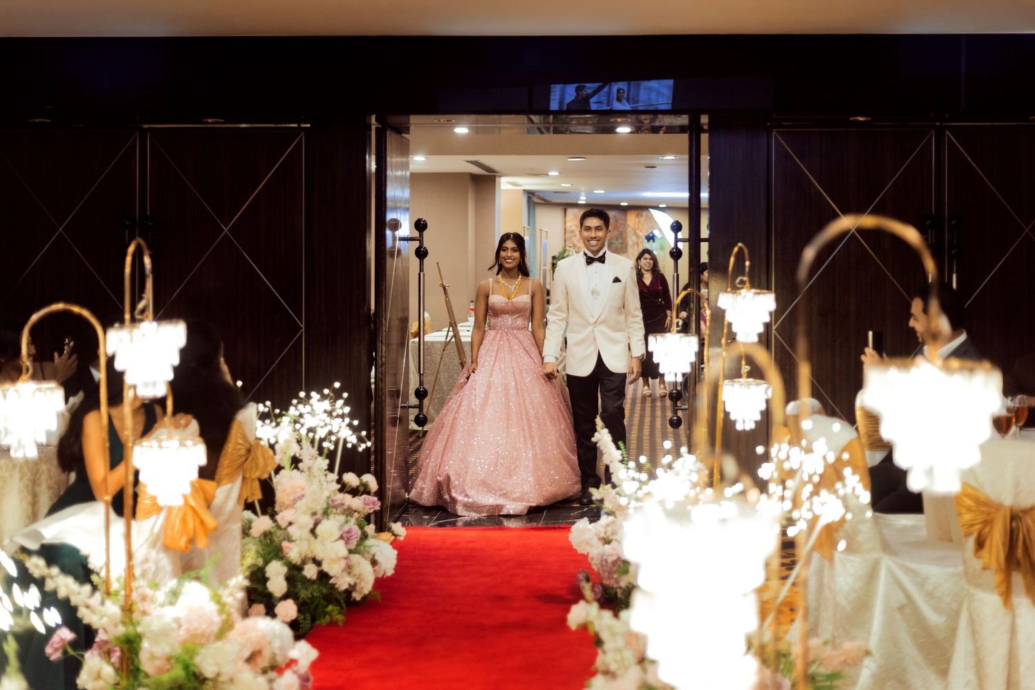Bride in pink ball gown and groom in white suit walk down red carpet aisle at elegant wedding reception venue.
