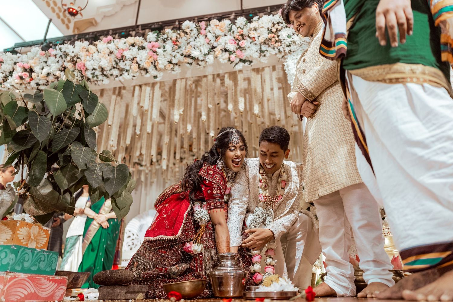 Bride and groom laughing during traditional Indian wedding ceremony with floral backdrop and ritual pot.