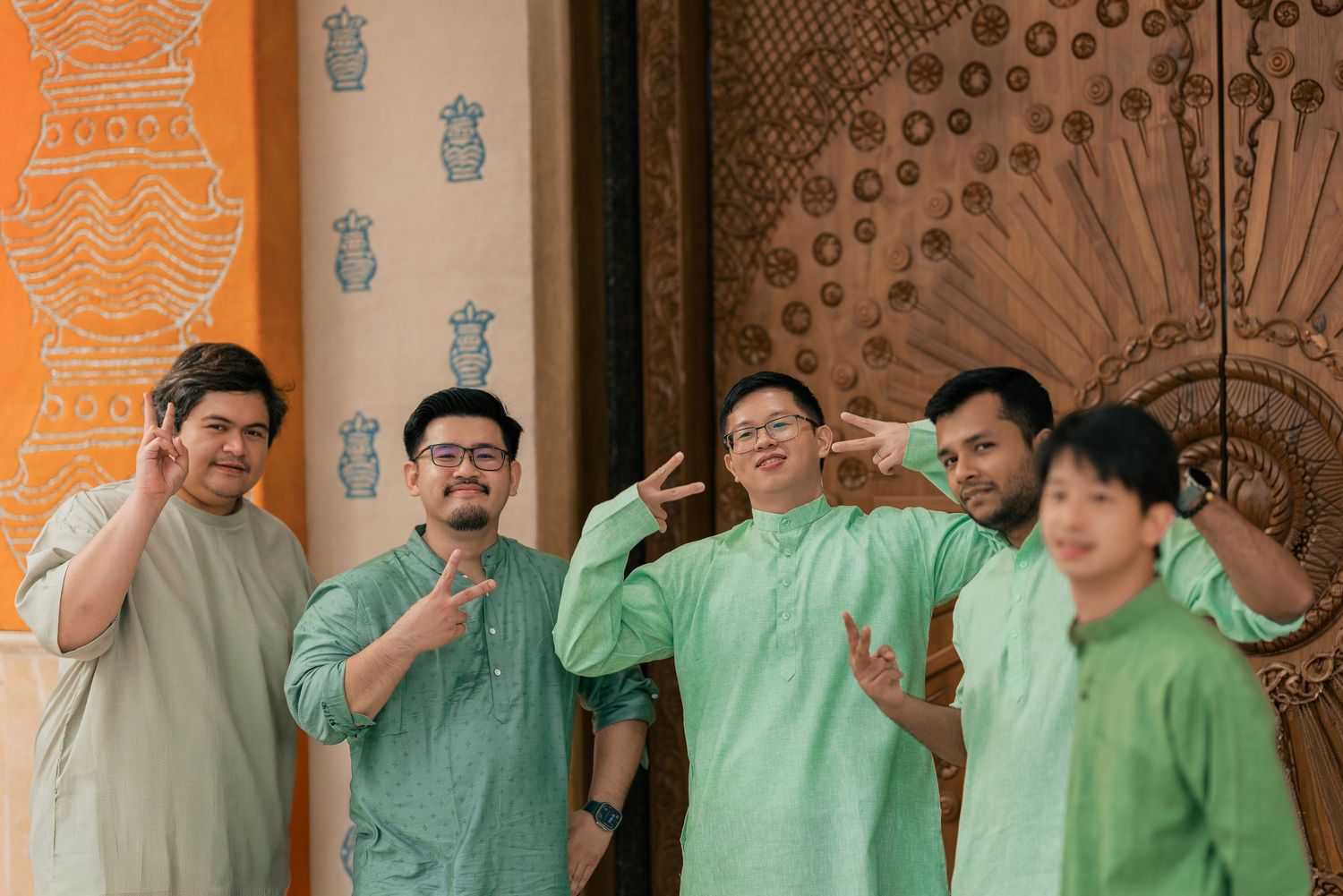 Five men in matching green traditional outfits smiling and posing together in front of ornate decor.