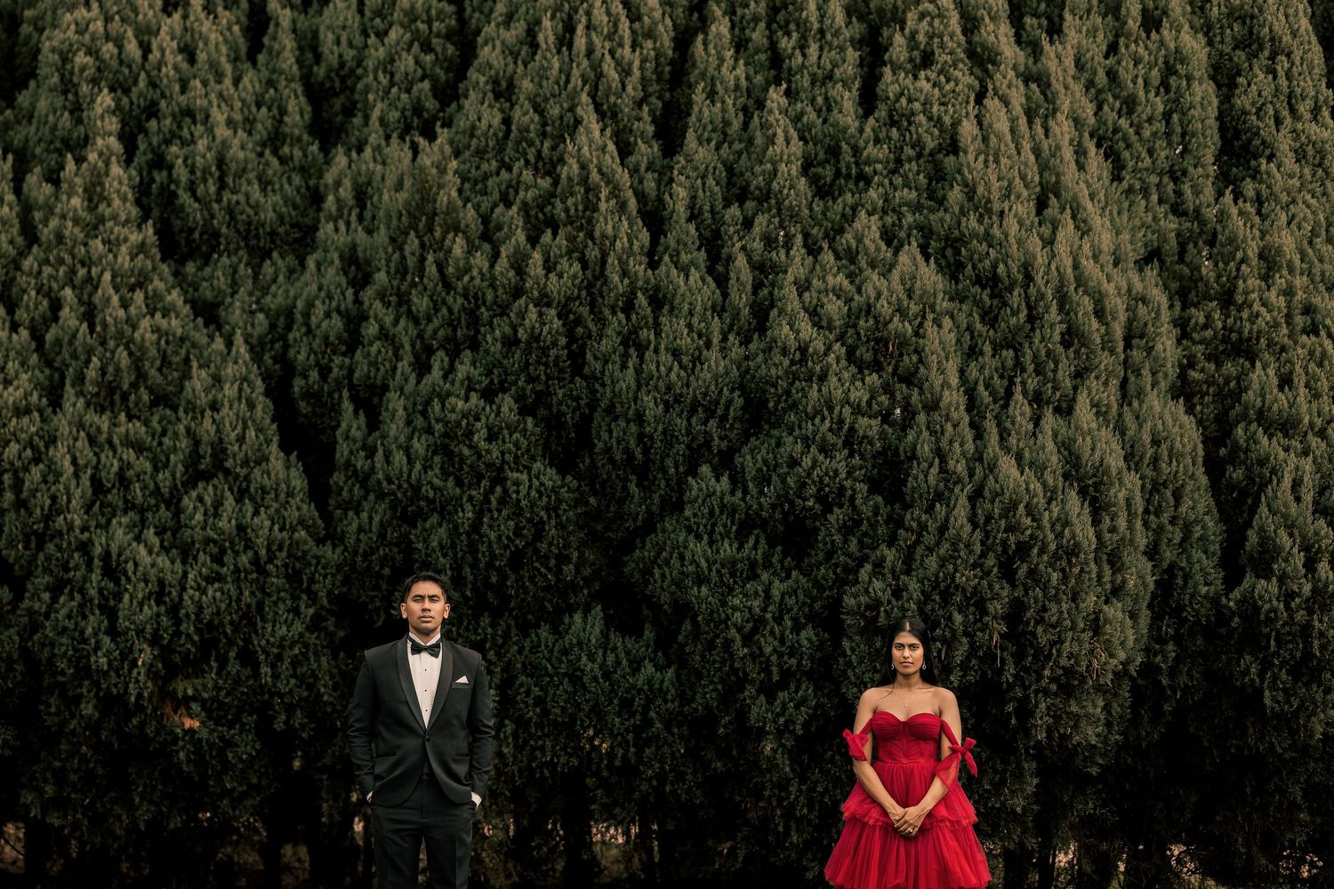 Couple in formal attire stand apart before tall green trees, man in black suit, woman in red ball gown.