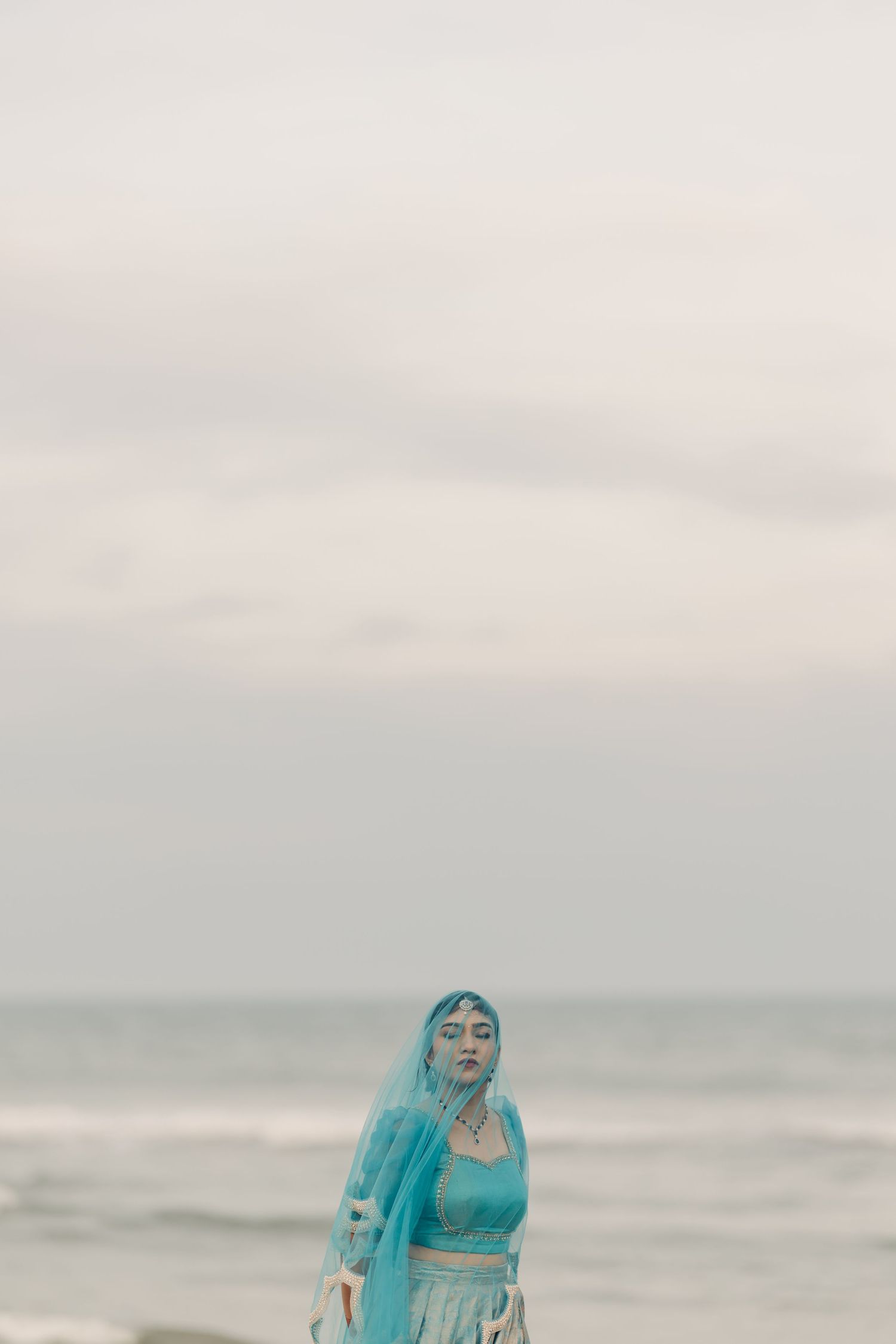 A figure in flowing turquoise fabric stands on a misty beach against an overcast sky.