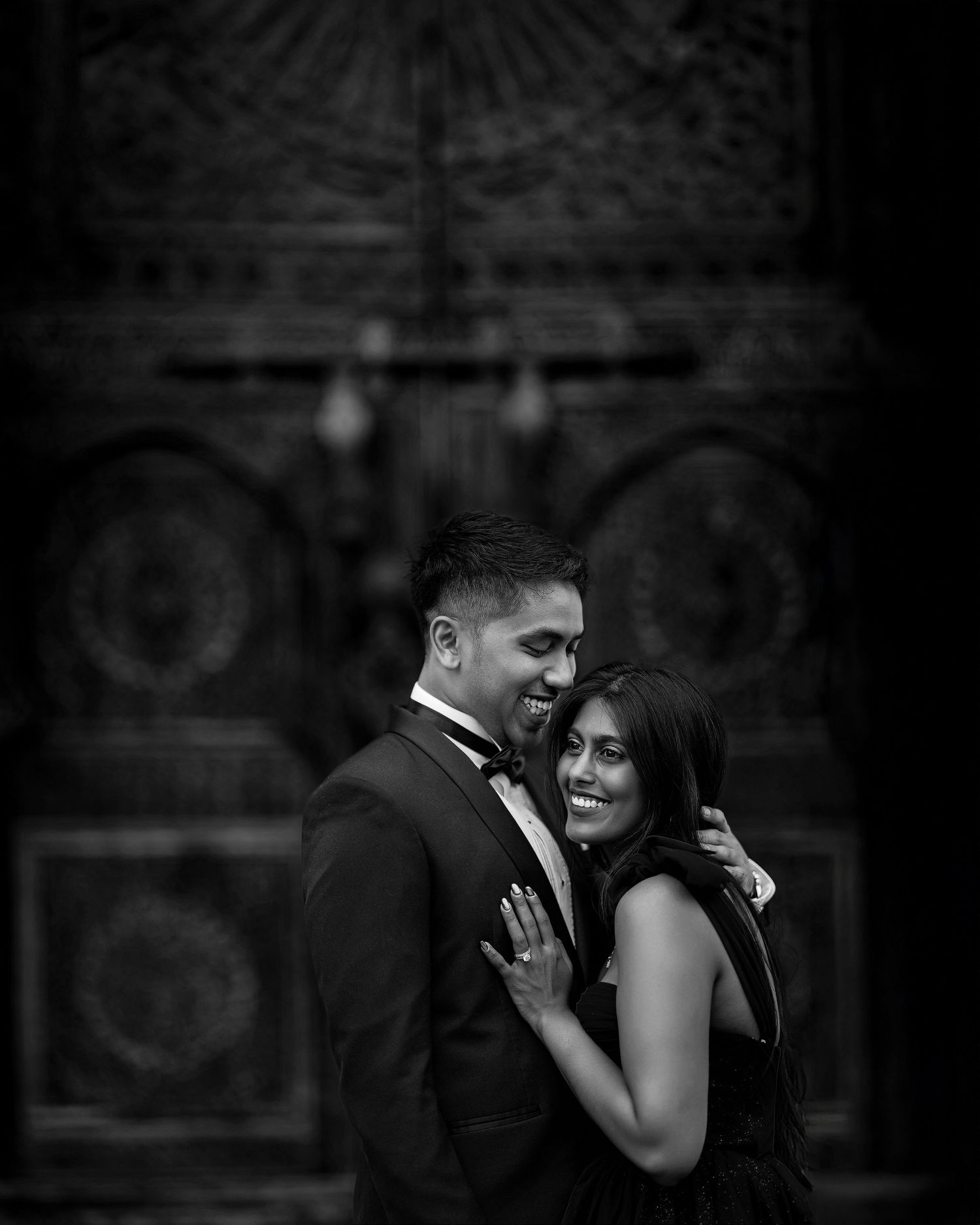 A romantic black and white portrait shows a couple embracing in front of an ornate architectural backdrop.