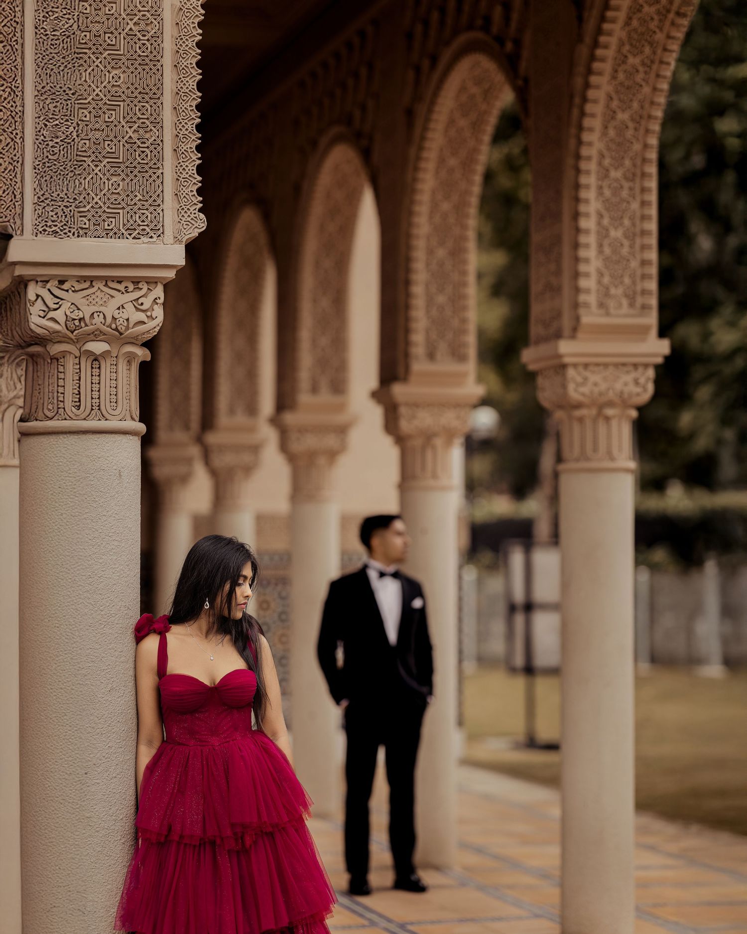 A couple in formal attire poses under ornate arched columns at a venue with intricate architectural details.