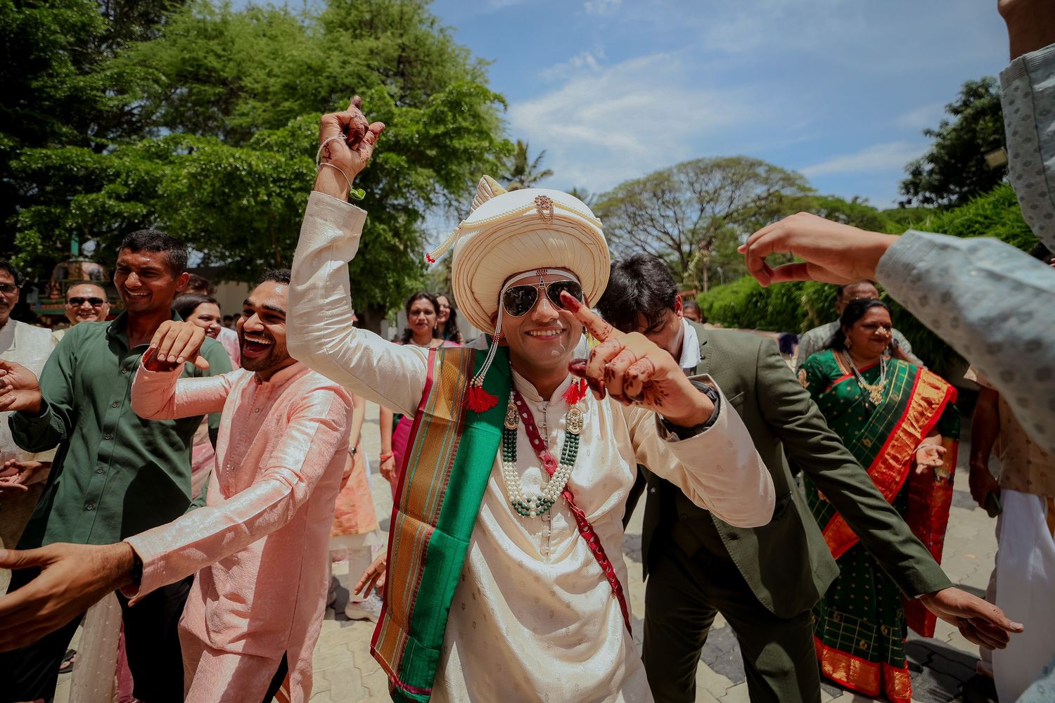 Wedding party celebrates outdoors with joyful dancing and raised arms in a garden setting.