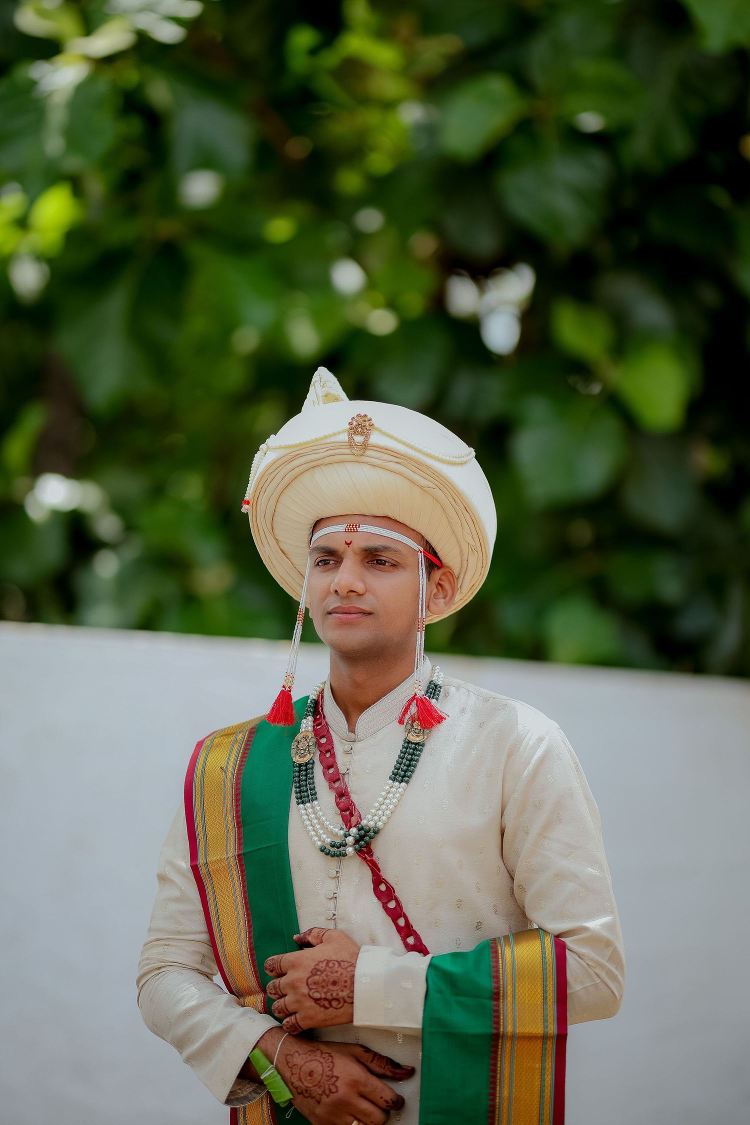 Traditional Indian groom wearing cream sherwani with green and gold sash poses for wedding portraits in outdoor setting.